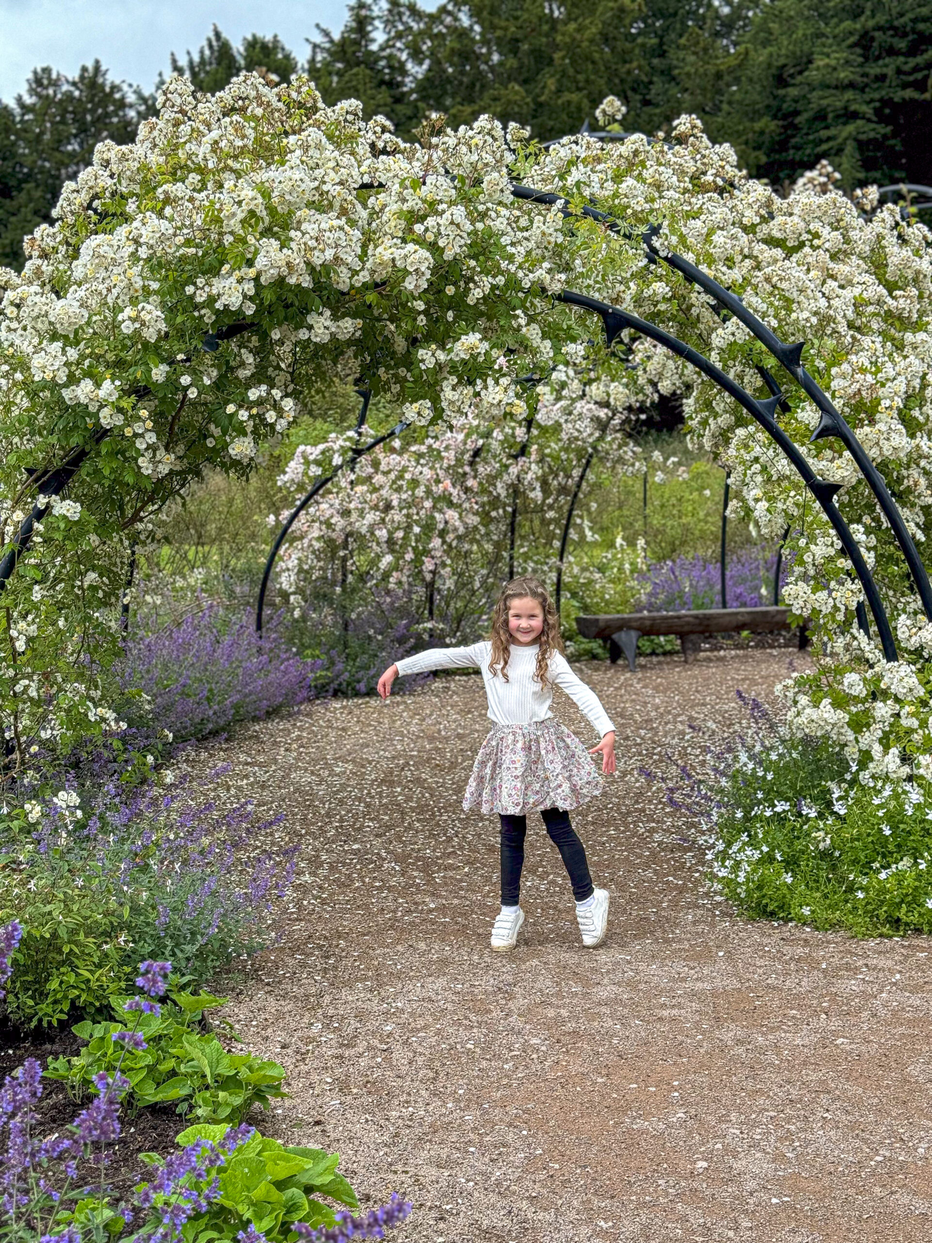 A smiling girl poses under a blooming flower arch in a garden surrounded by greenery and purple flowers.