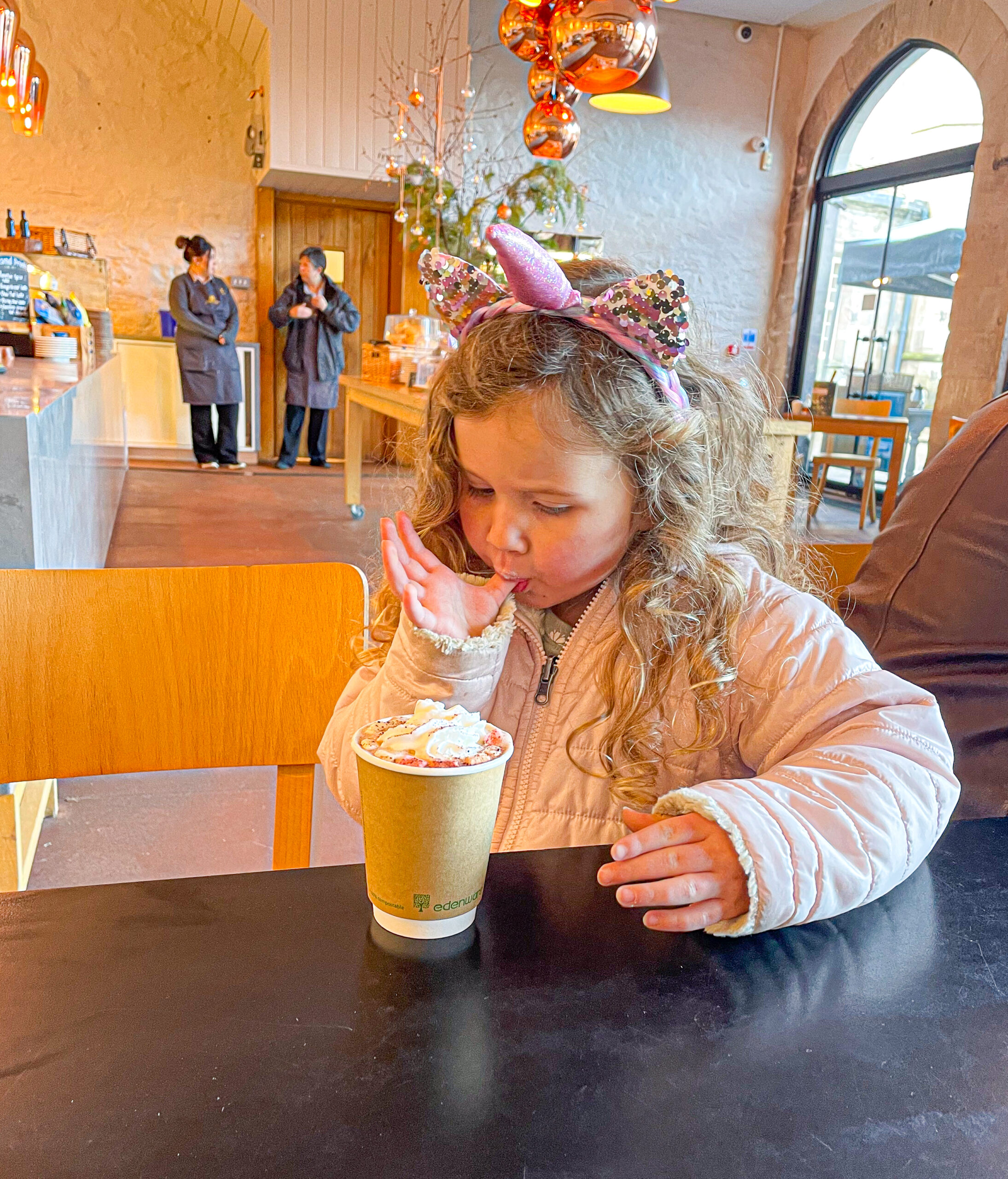 Young girl wearing a unicorn headband tastes whipped cream from her hot drink at a cozy cafe.