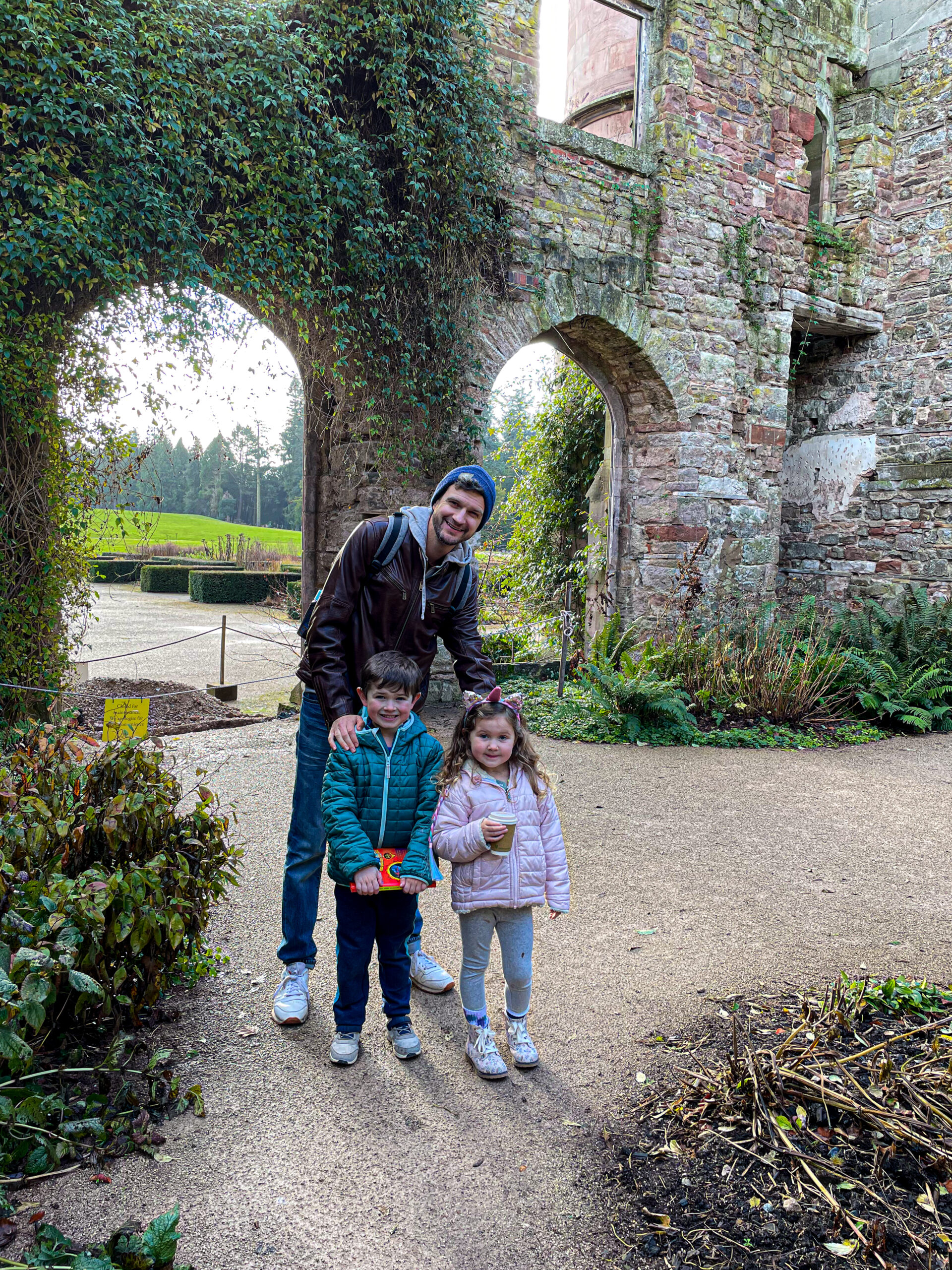 A man and two children smiling, standing outside near old stone ruins and greenery.