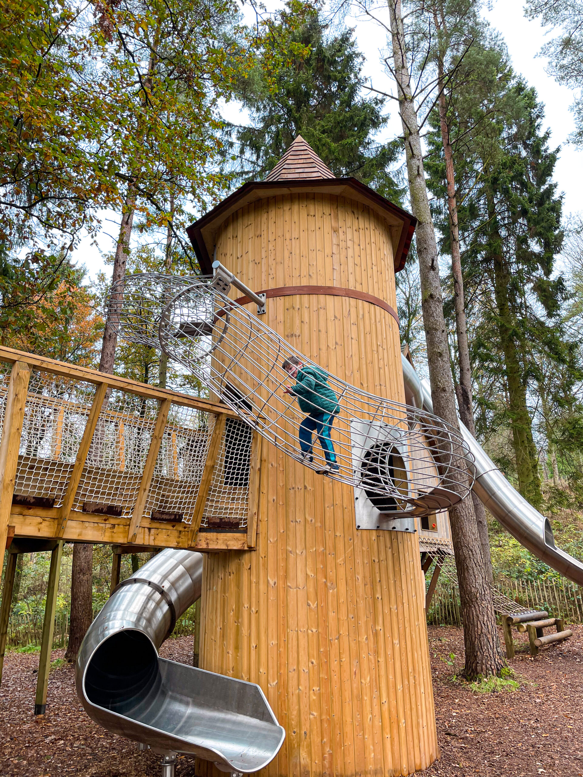 Child climbing a metal tunnel at a wooden playground tower surrounded by tall trees and slides.