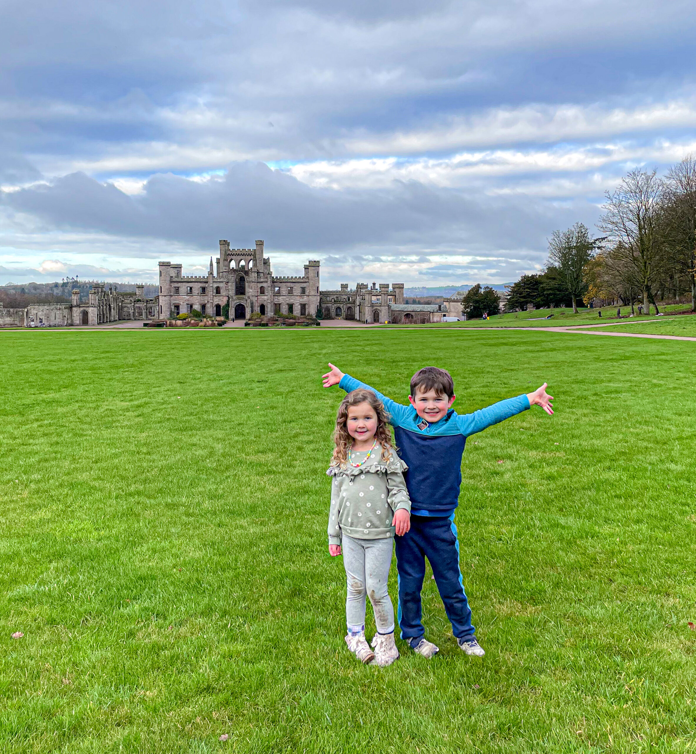 Two children stand on green grass with a historic castle building in the background under a cloudy sky.