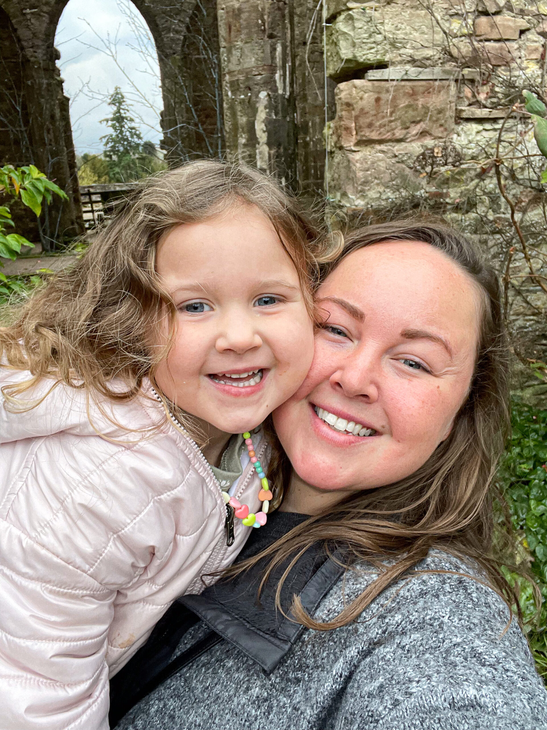 Smiling woman and young girl outdoors, posing for a selfie in front of old stone ruins and greenery.