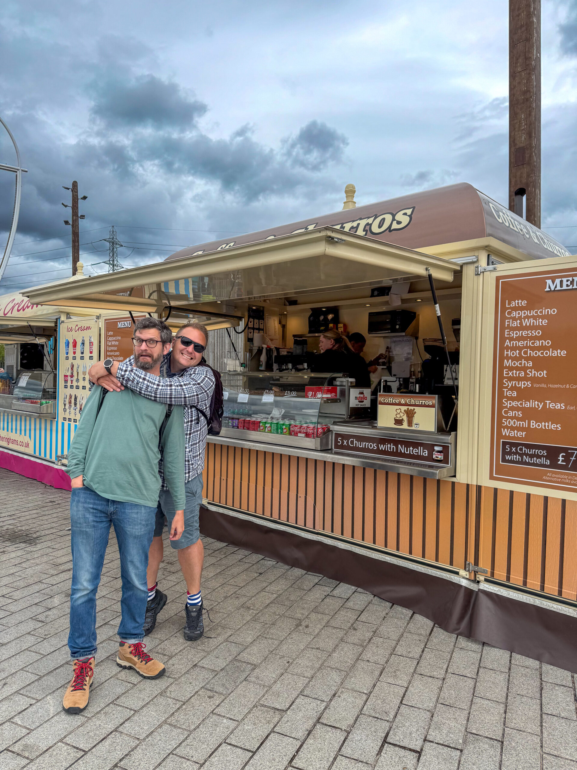 Two men pose in front of a churros food stall; one hugs the other from behind on a cloudy day.
