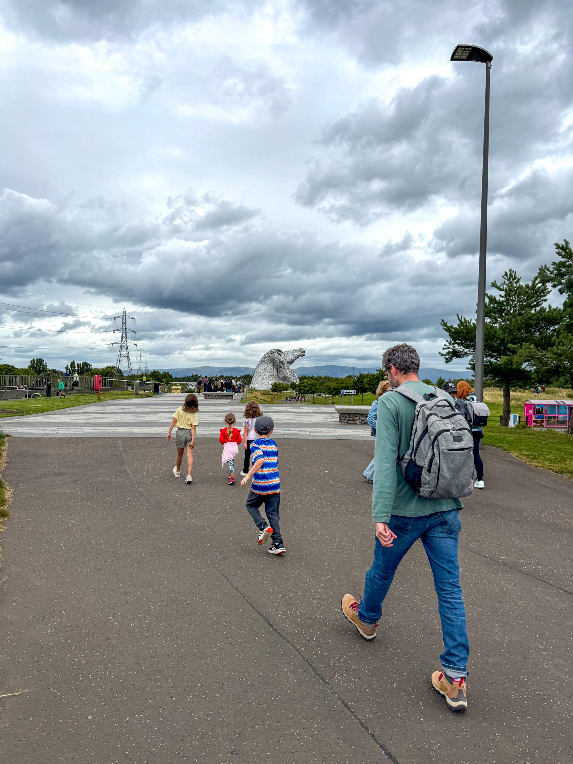 A man and children walk on a path toward a large horse-head sculpture under a cloudy sky.