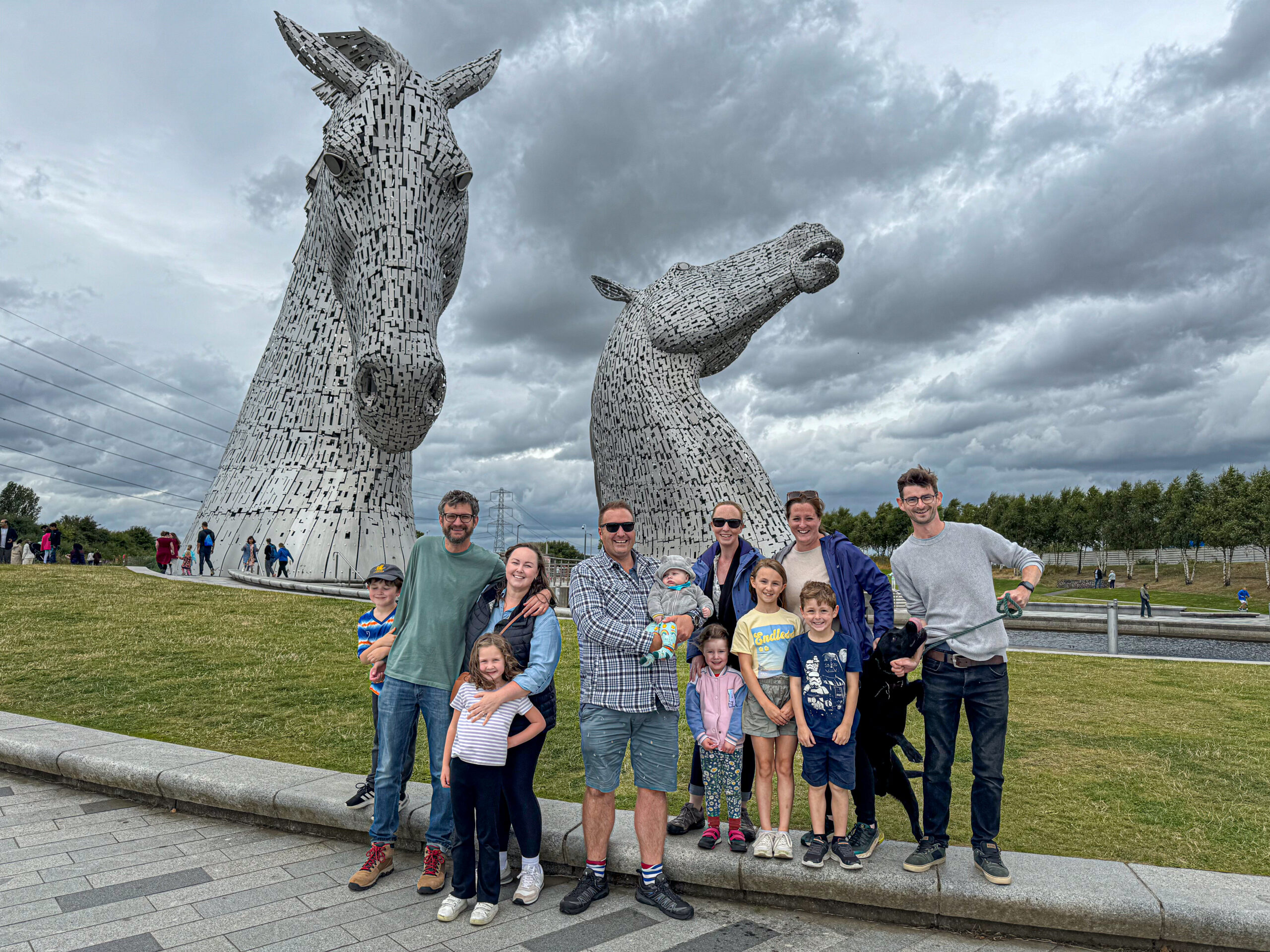 A group of people pose in front of The Kelpies horse-head sculptures under a cloudy sky.