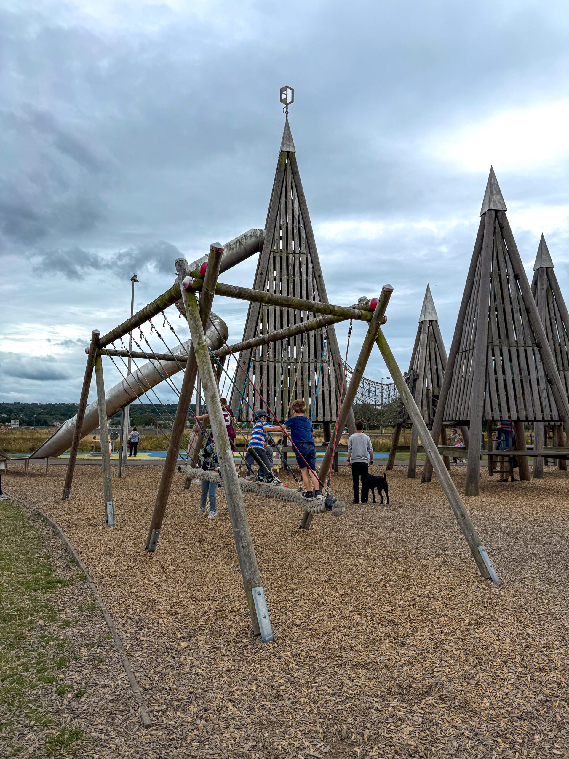Children play on a rope swing and climb wooden structures at an outdoor playground under a cloudy sky.