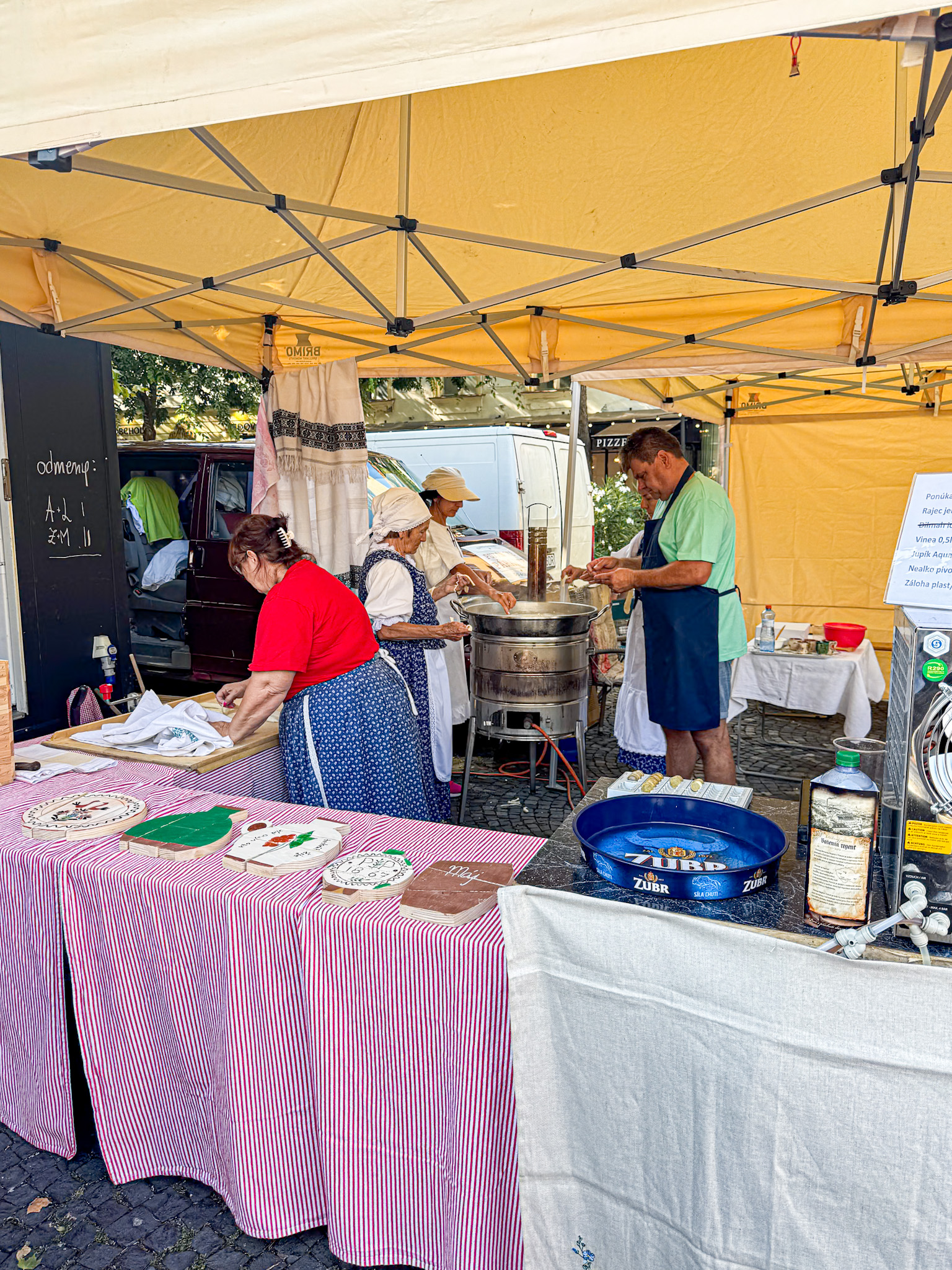 Three people in aprons prepare food at a market stall under a yellow canopy with various items on display.