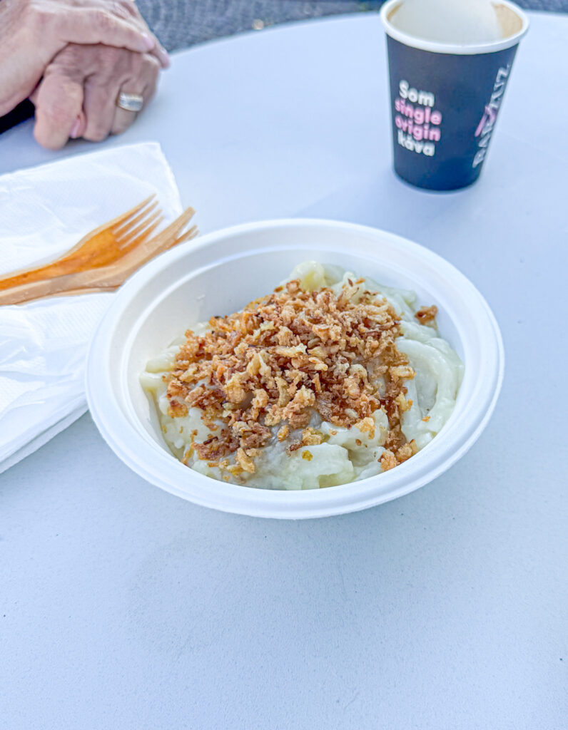A bowl of dumplings topped with fried onions on a table, near a napkin, fork, and a paper cup.