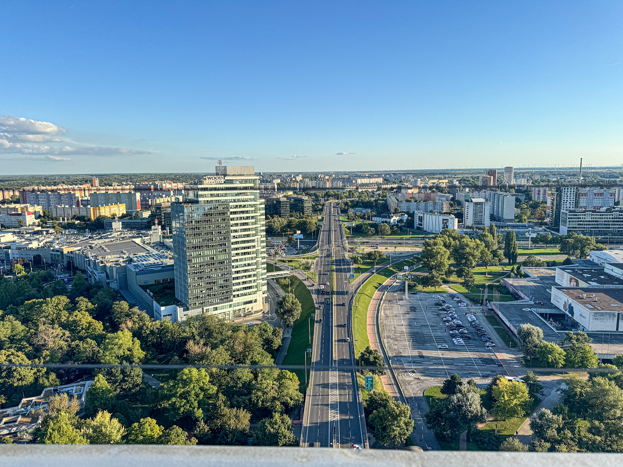 Aerial view of a cityscape with tall buildings, roads, parked cars, and green trees under a clear blue sky.