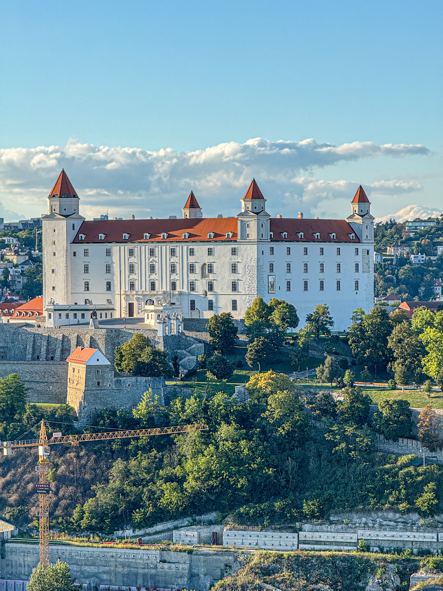 Bratislava castle with red roofs and four towers, surrounded by trees and a city in the background.