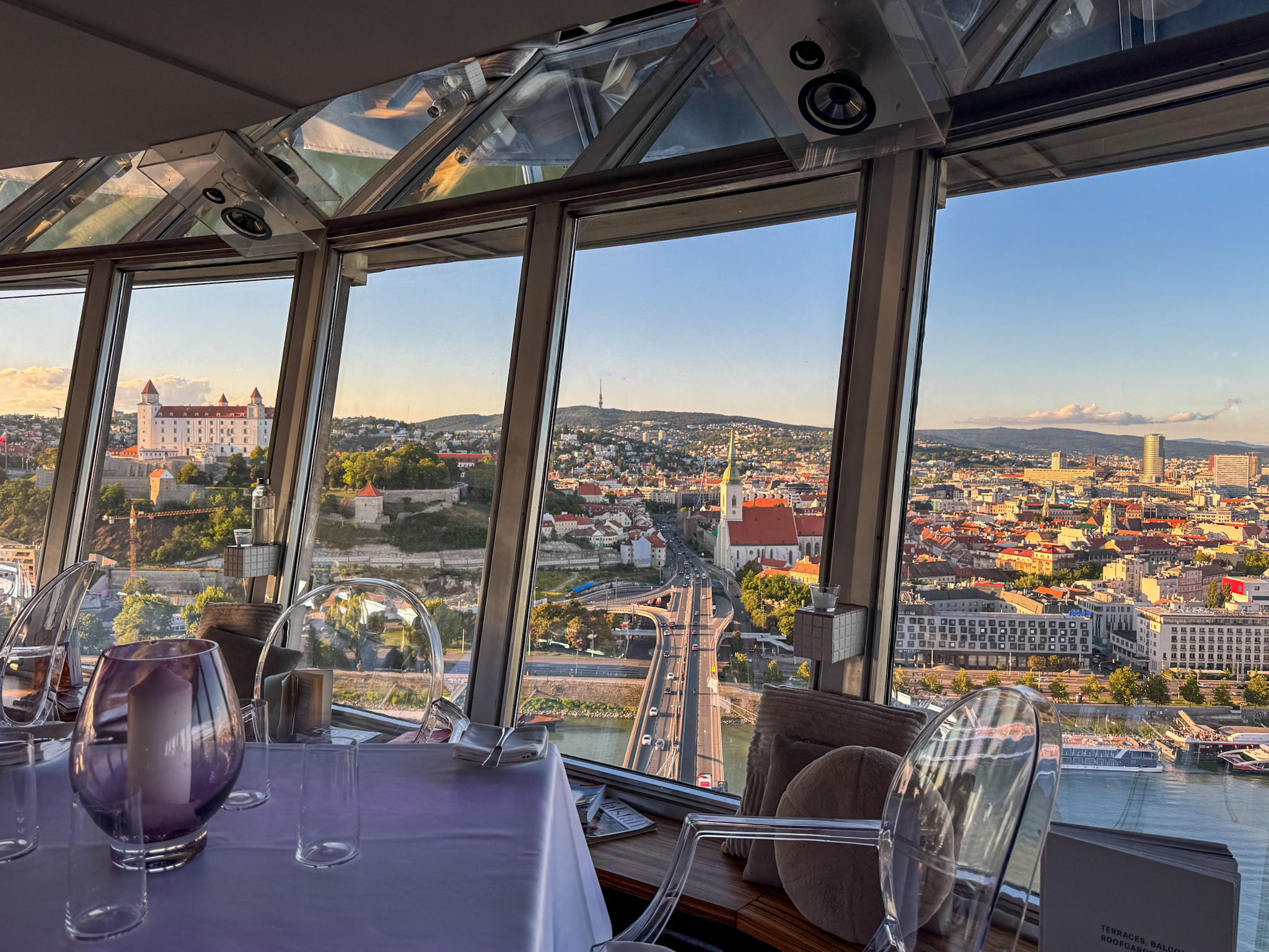 Table set for dining at the UFO Tower Restaurant by large windows overlooking the Bratislava cityscape with a bridge and historic buildings at sunset.