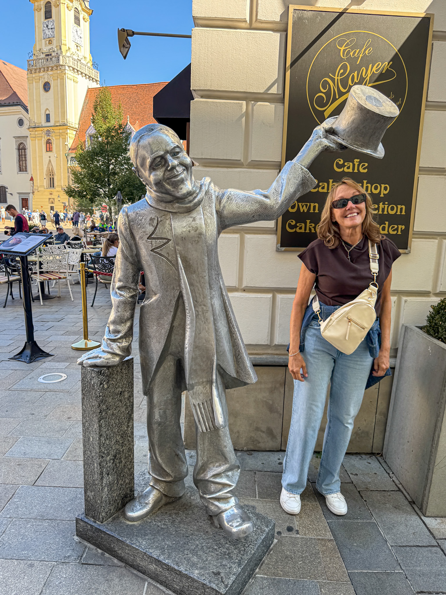Smiling woman stands next to a silver painted statue tipping its hat on Bratislava sidewalk near a cafe.