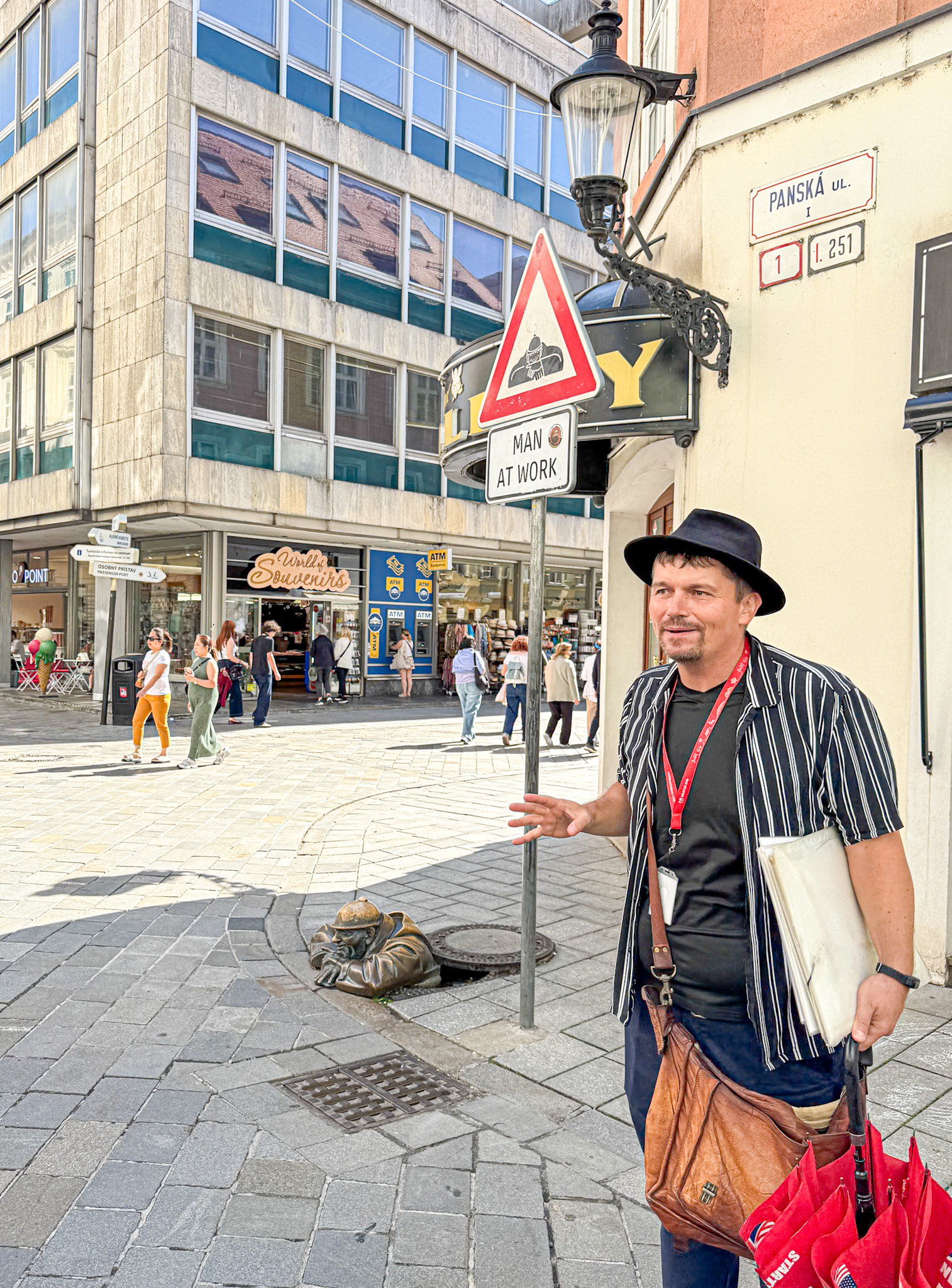 A man stands by a Man at Work sign next to a bronze statue emerging from a street manhole.