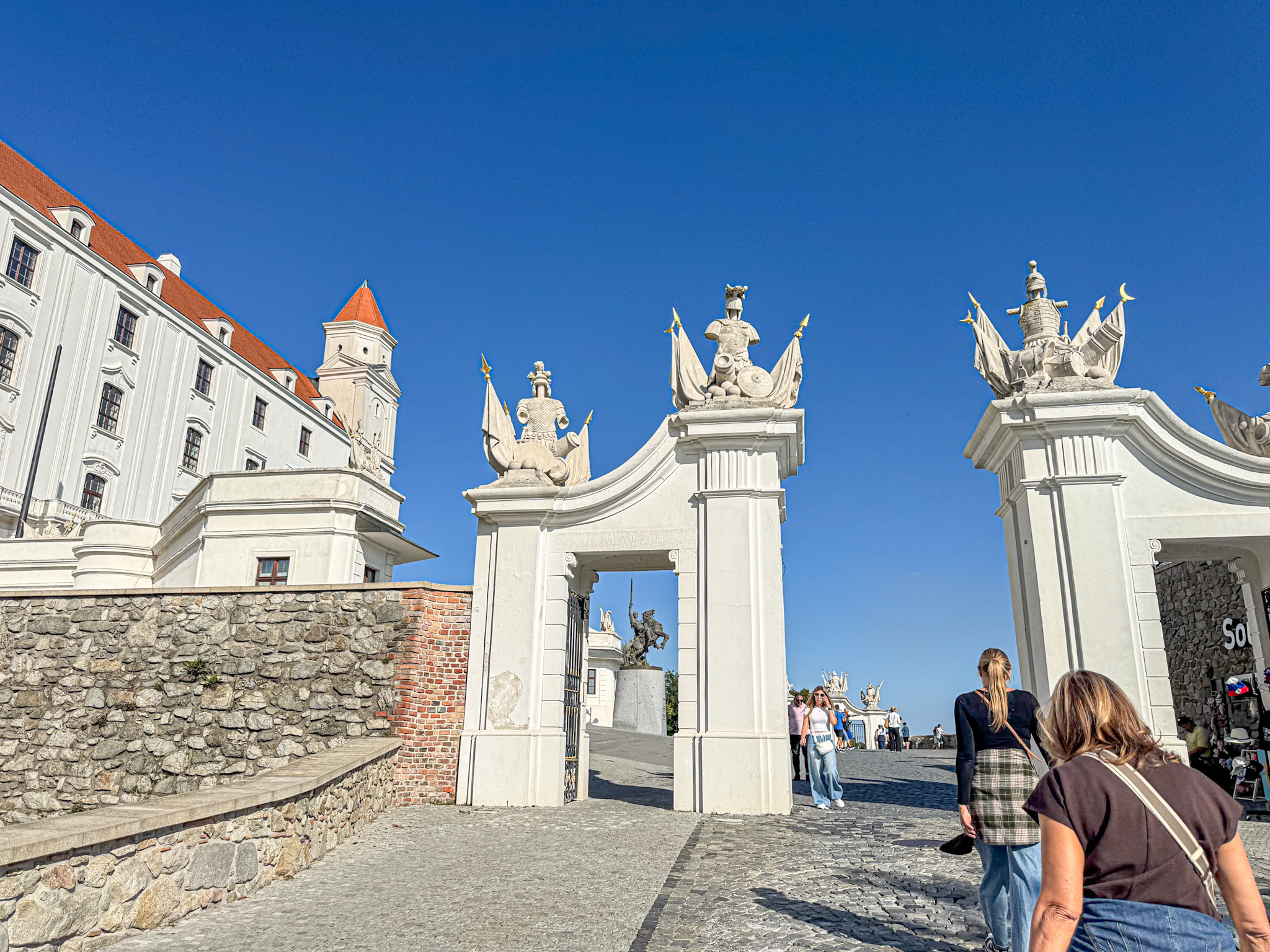 People walk through ornate white gates beside Bratislava castle under a clear blue sky.