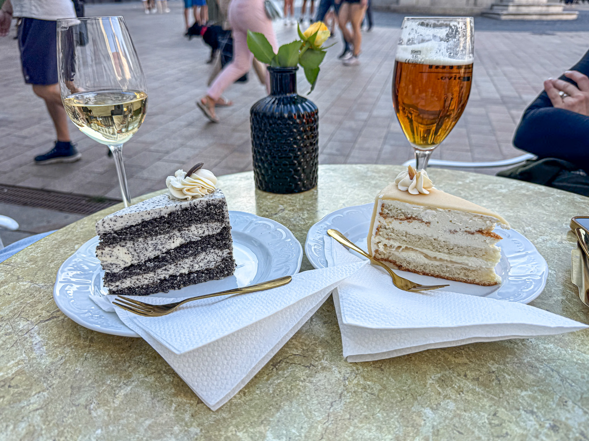 Two slices of cake, a glass of white wine, and a beer on an outdoor café table with people walking by.