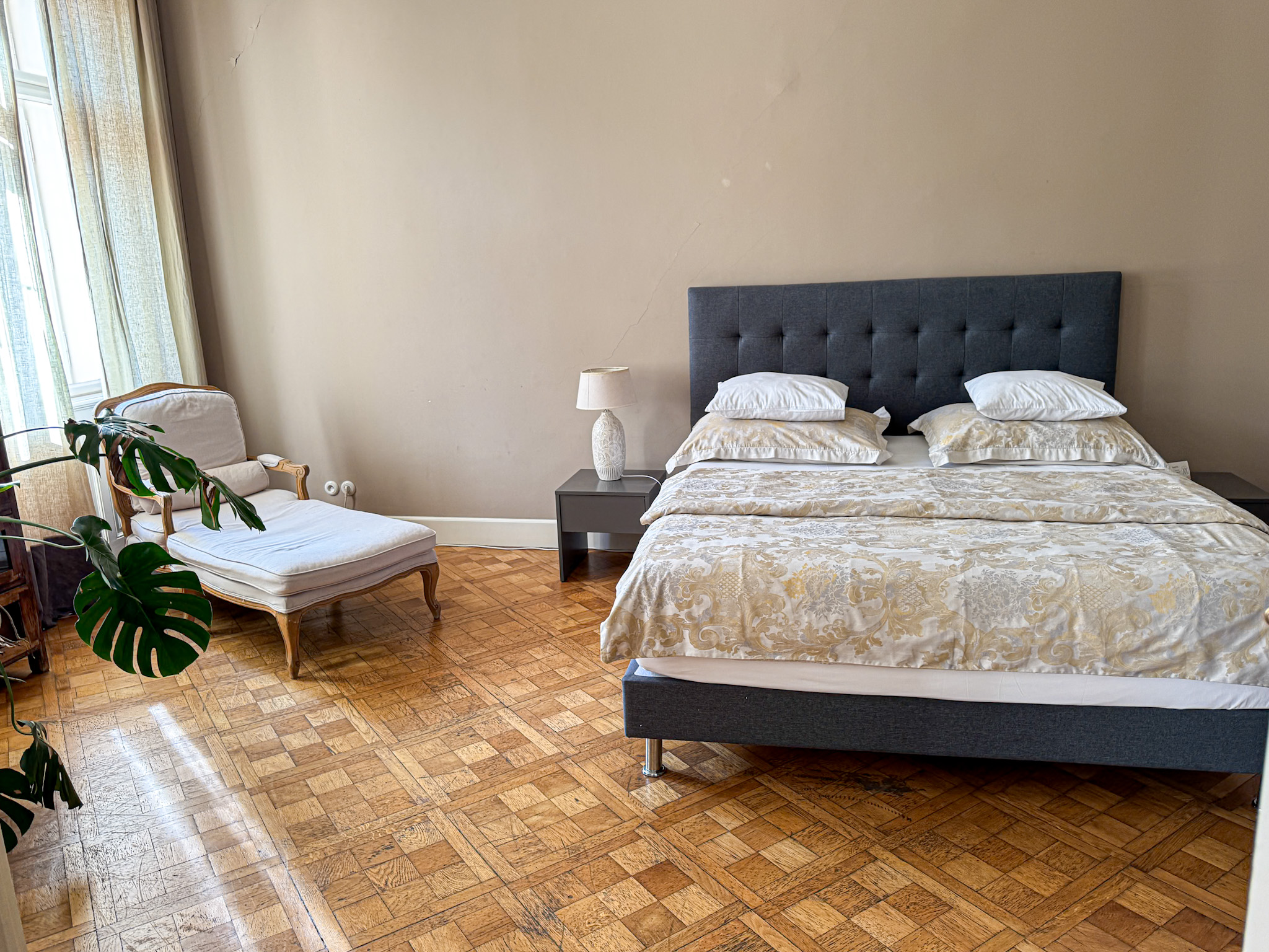 Modern hotel bedroom with a gray bed, patterned bedding, a white armchair, and a leafy plant on parquet floor at the Malostranská Residence in Prague, Czech Republic.