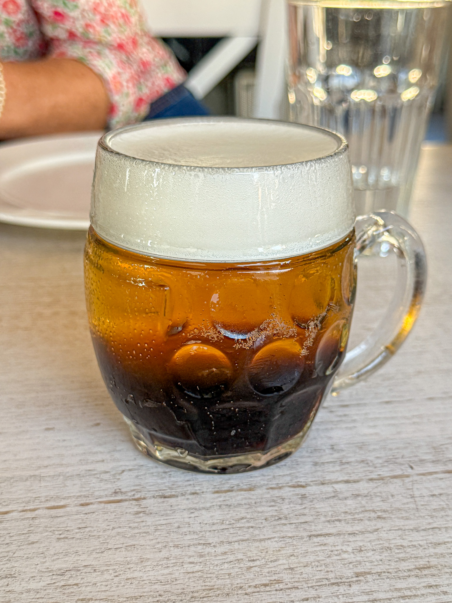 A glass mug of dark and light layered beer sits on a table with a plate and glass in the background.