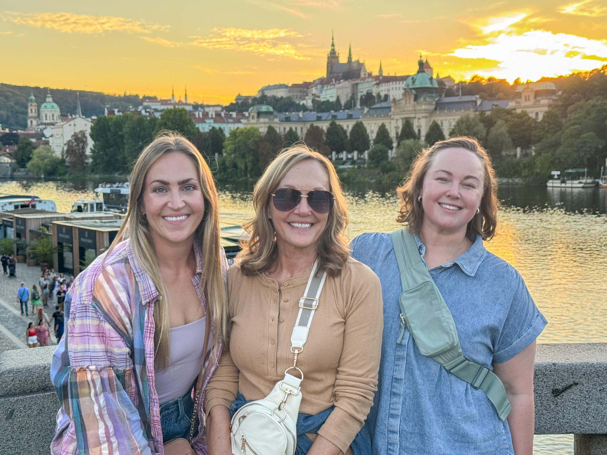 Three women smiling on a bridge at sunset with historic buildings and a river in the background.