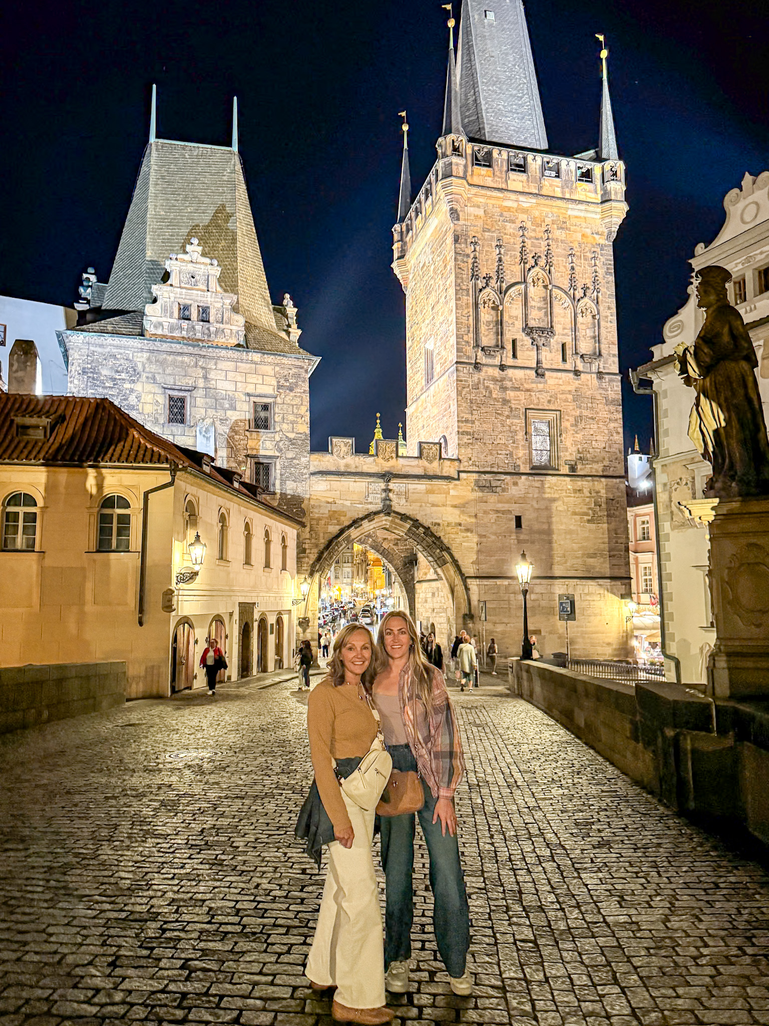 Two women pose and smile on a cobblestone street at night near Charles Bridge in Prague.