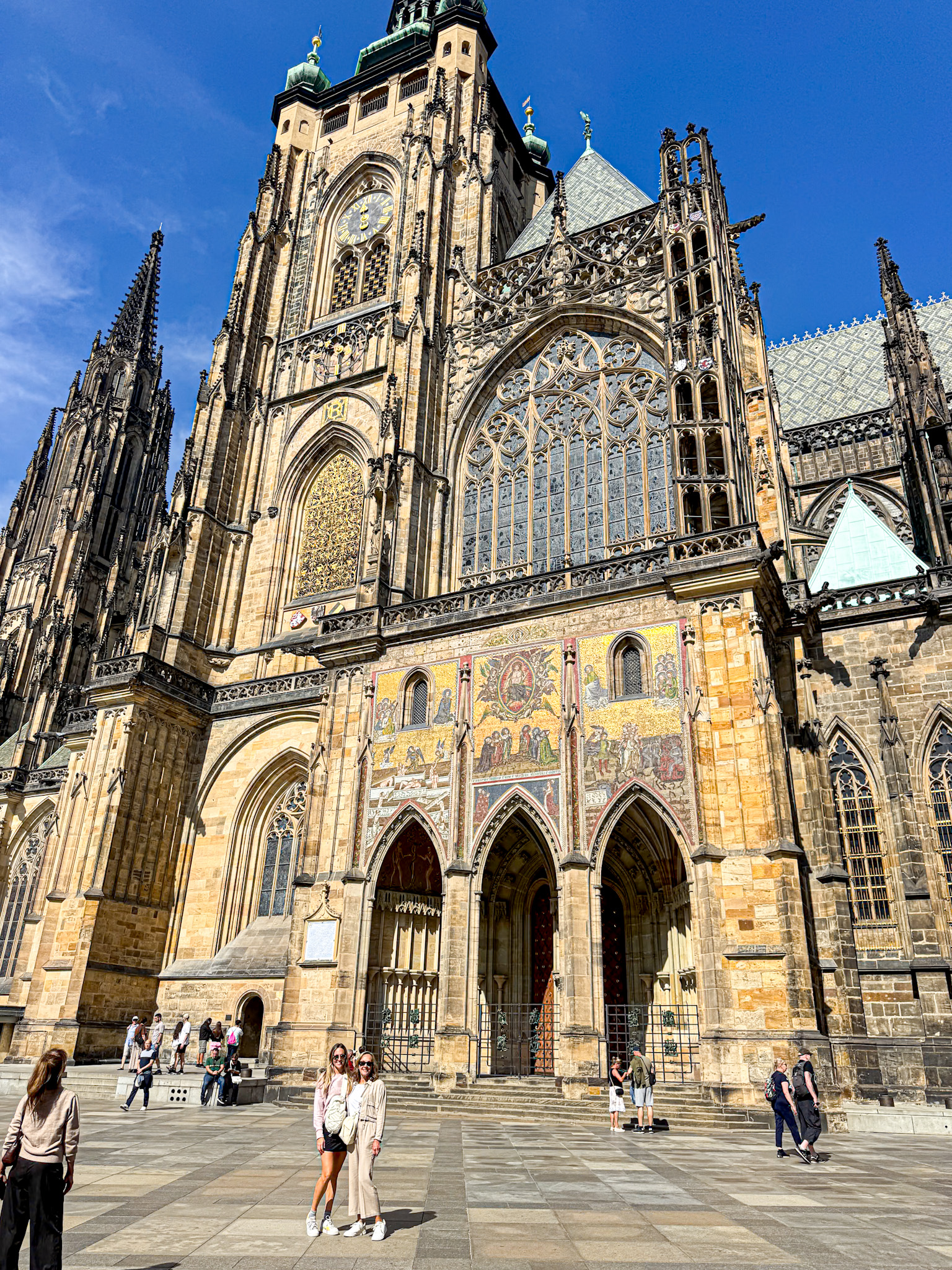 People stand in front of a large, ornate a Prague Gothic cathedral with intricate details and stained glass windows.