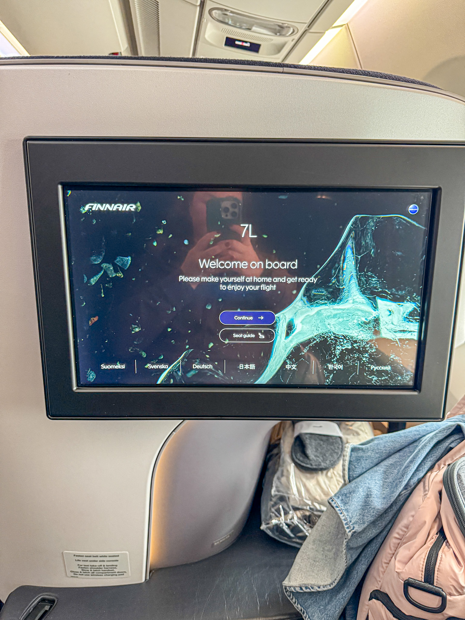 Seatback screen on a Finnair flight displays a welcome message and seat number 7L, with bags below the screen.