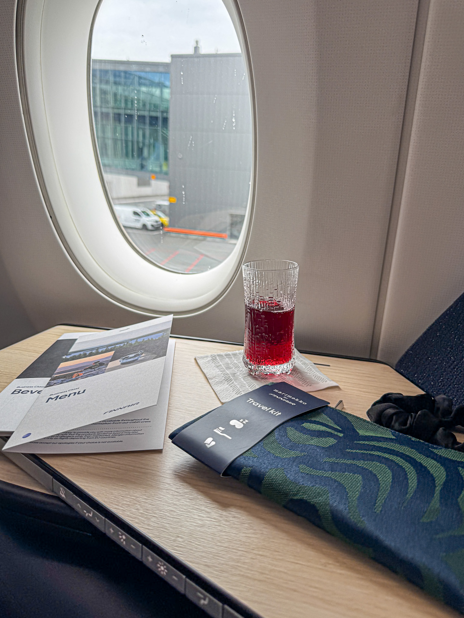 A glass of blueberry juice, menus, and an amenity kit on a Finnair business class airplane tray by a window at the airport gate.