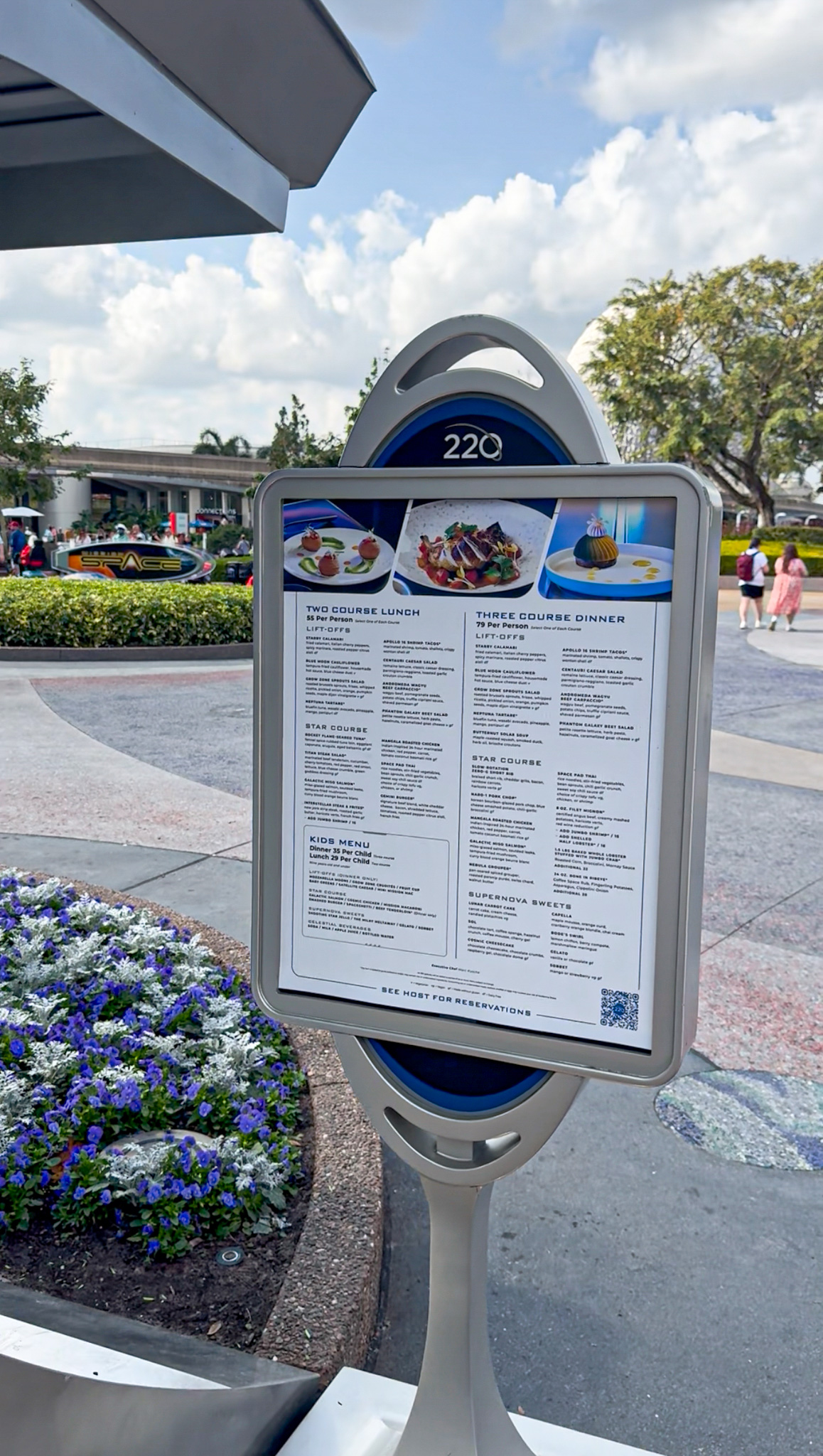 Outdoor Space 220 restaurant menu sign with lunch and dinner options, near flowers and people walking in the background.