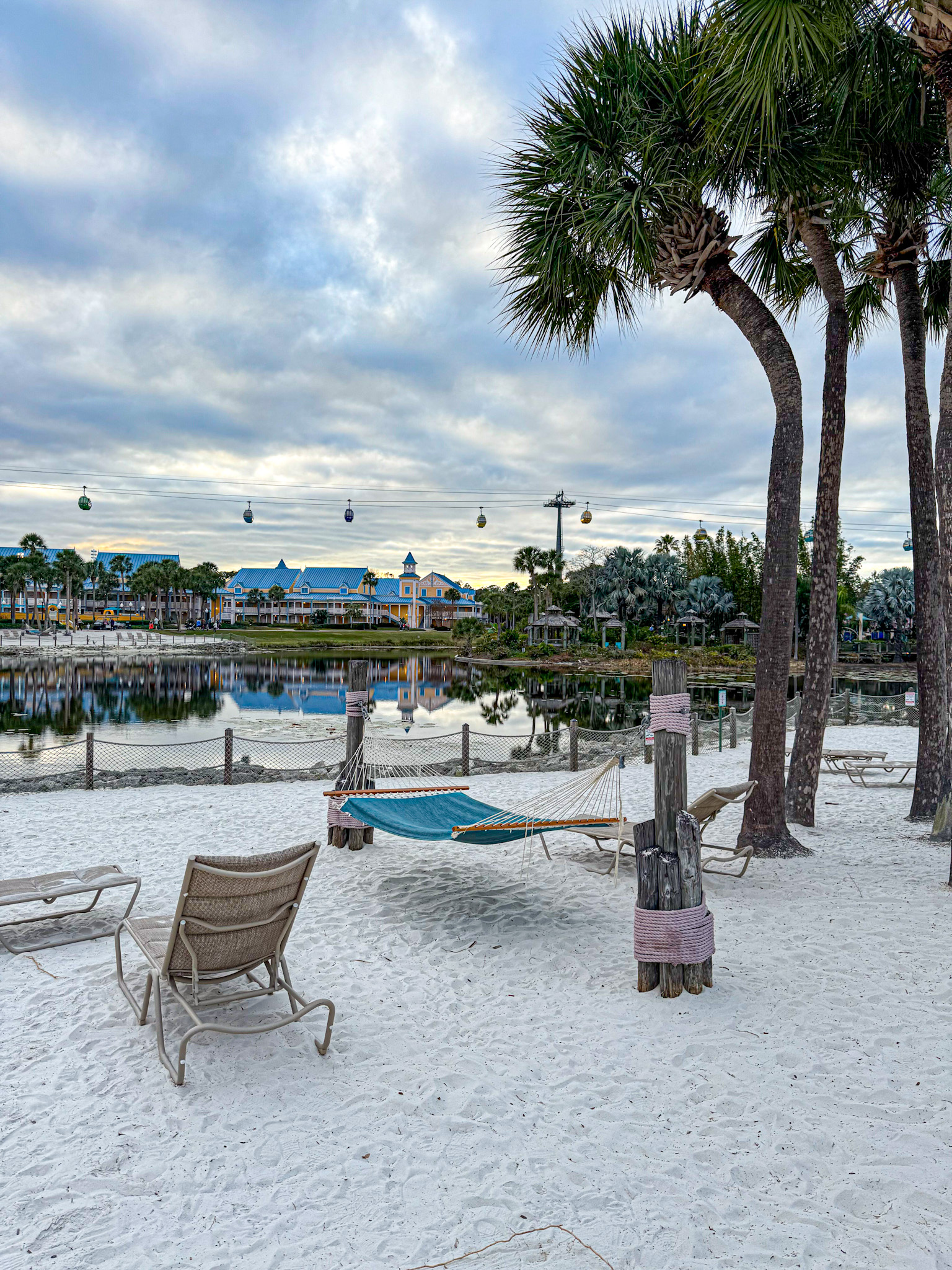 Hammock and lounge chairs on a sandy beach with palm trees by a calm lake, under a cloudy sky at Disney World.