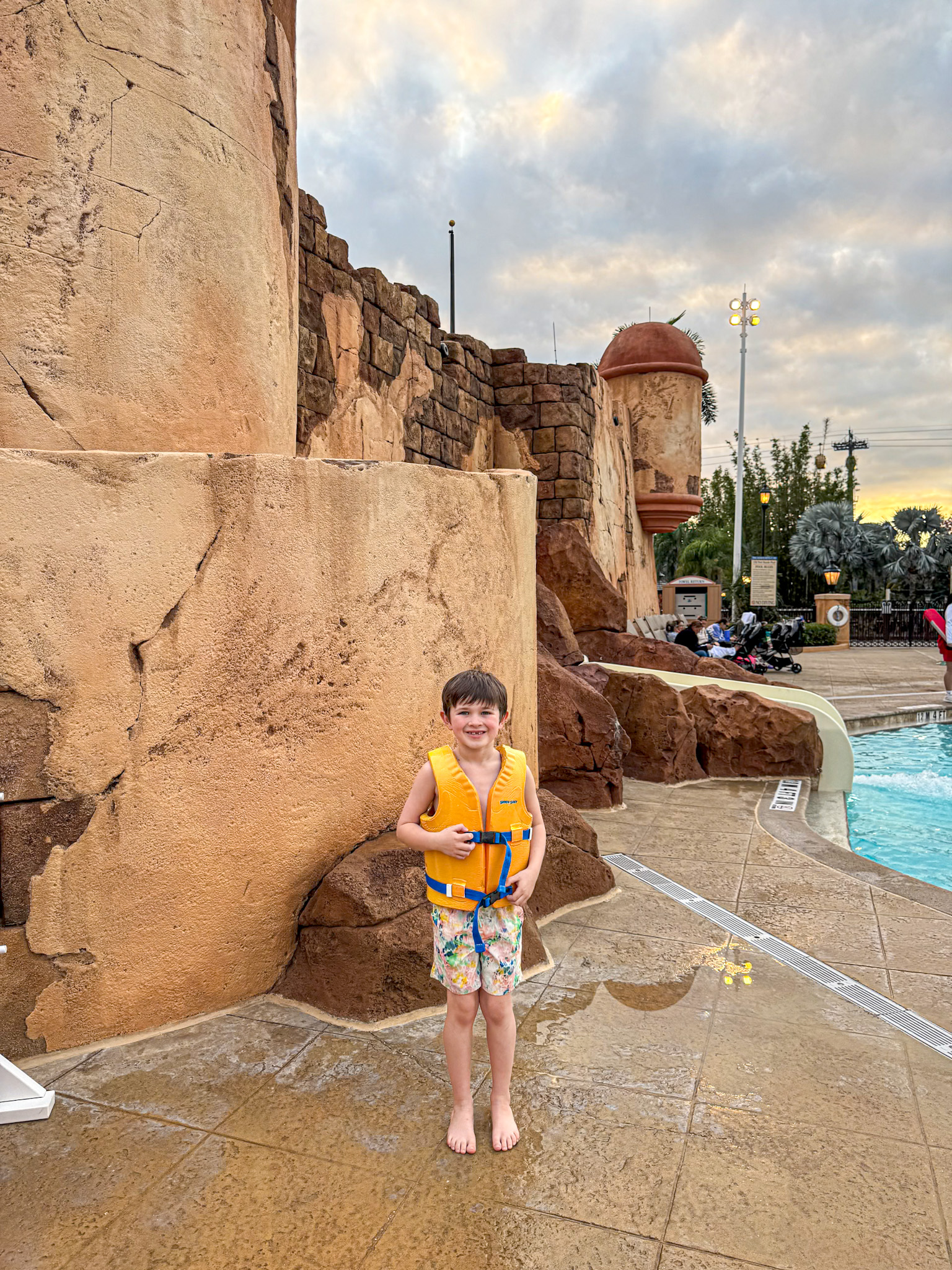 Young boy in a yellow life vest smiles by a pool at Disney's Caribbean Beach Resort, with stone walls and cloudy sky in the background.