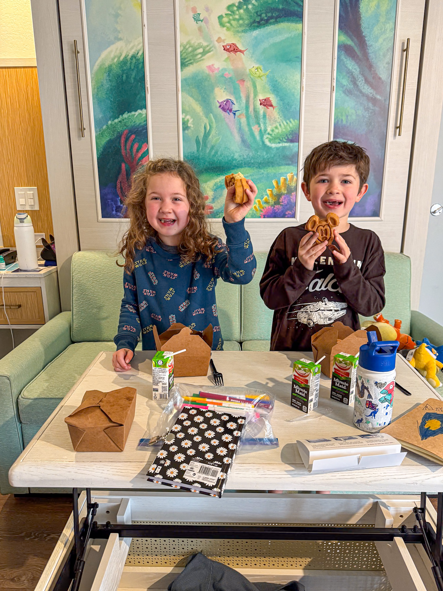 Two smiling children hold mickey waffles while standing by a table with food and drinks in a cozy living space at Caribbean Beach at Disney World.