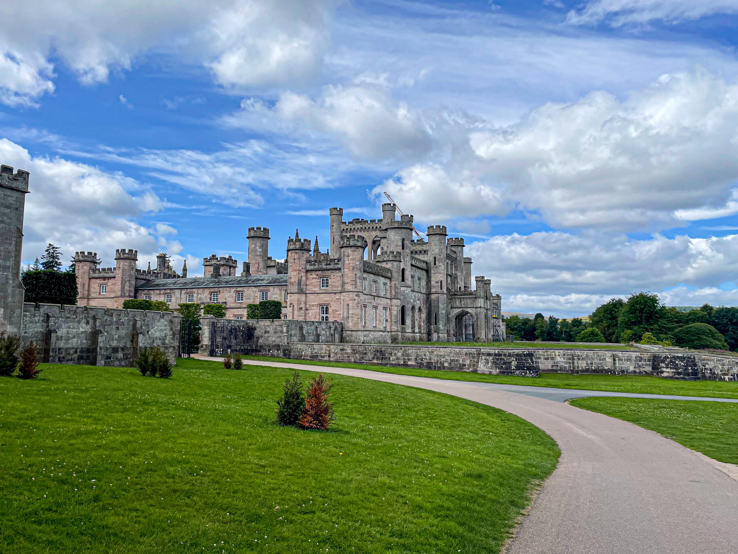 A large stone castle with towers and battlements sits behind a curved path and green lawn under a partly cloudy sky.