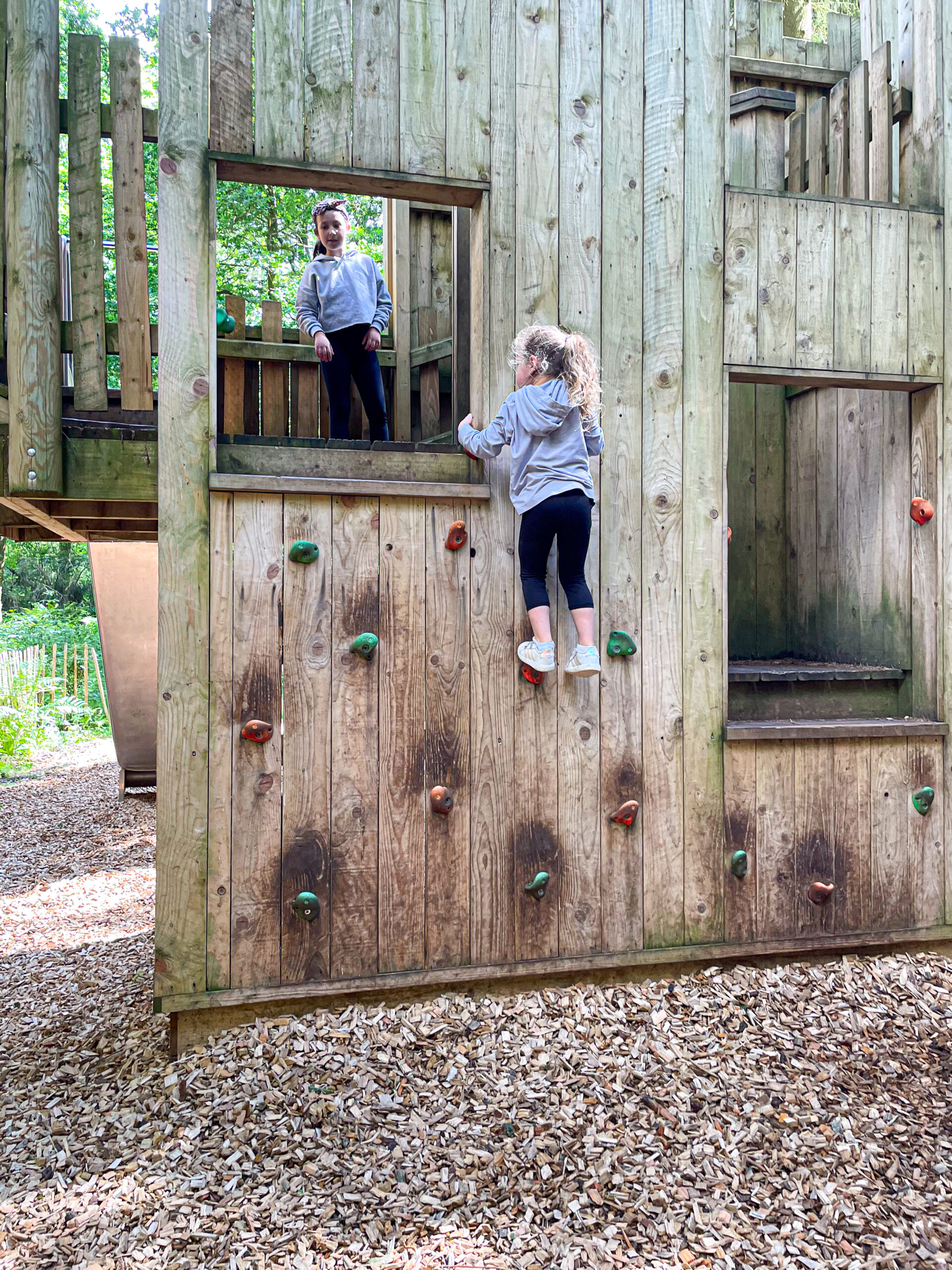 Two children play on a wooden climbing wall; one climbs while the other stands at a window above.