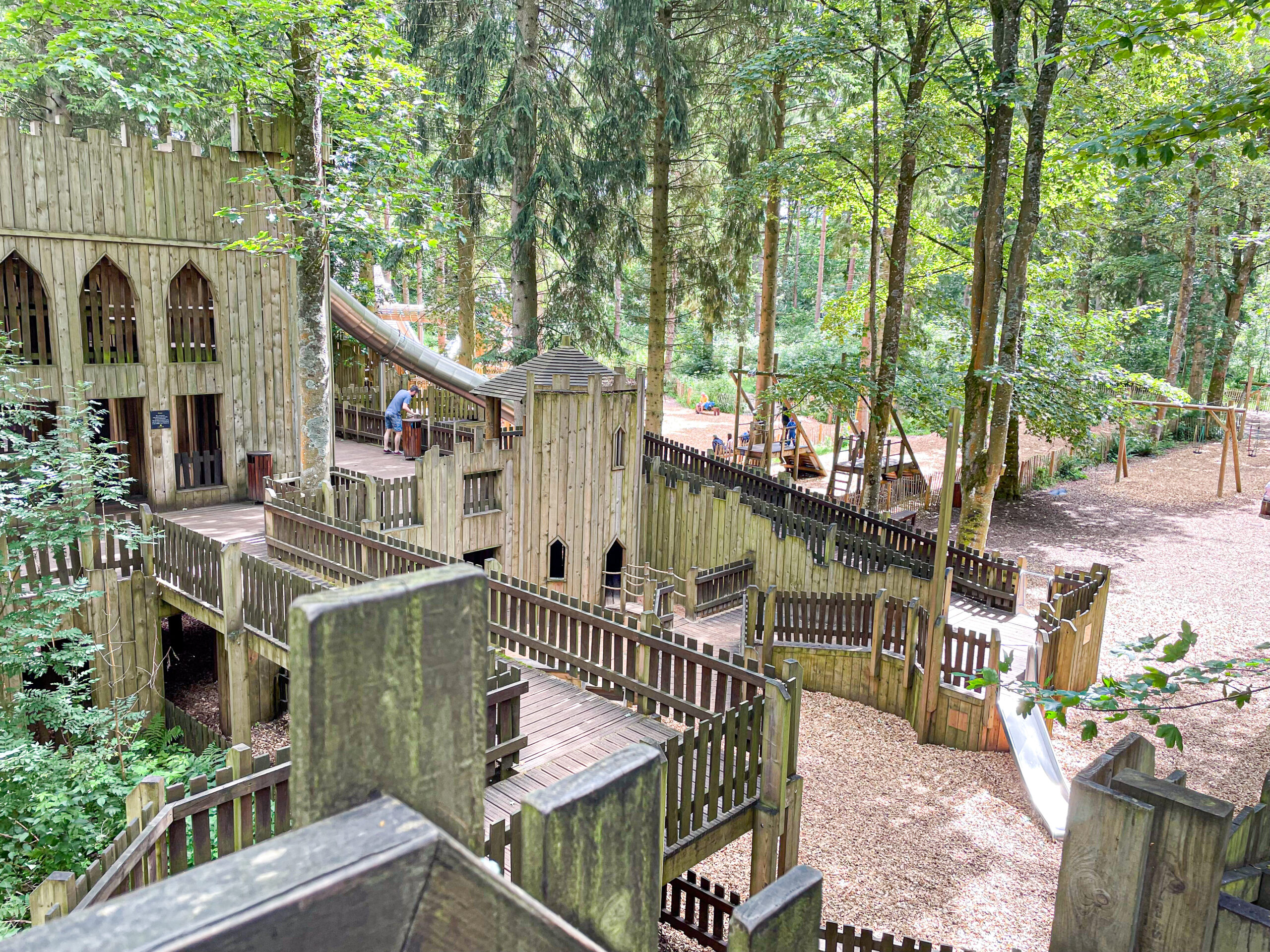 Wooden playground castle with towers, walkways, and slides set in a forested, tree-filled area.