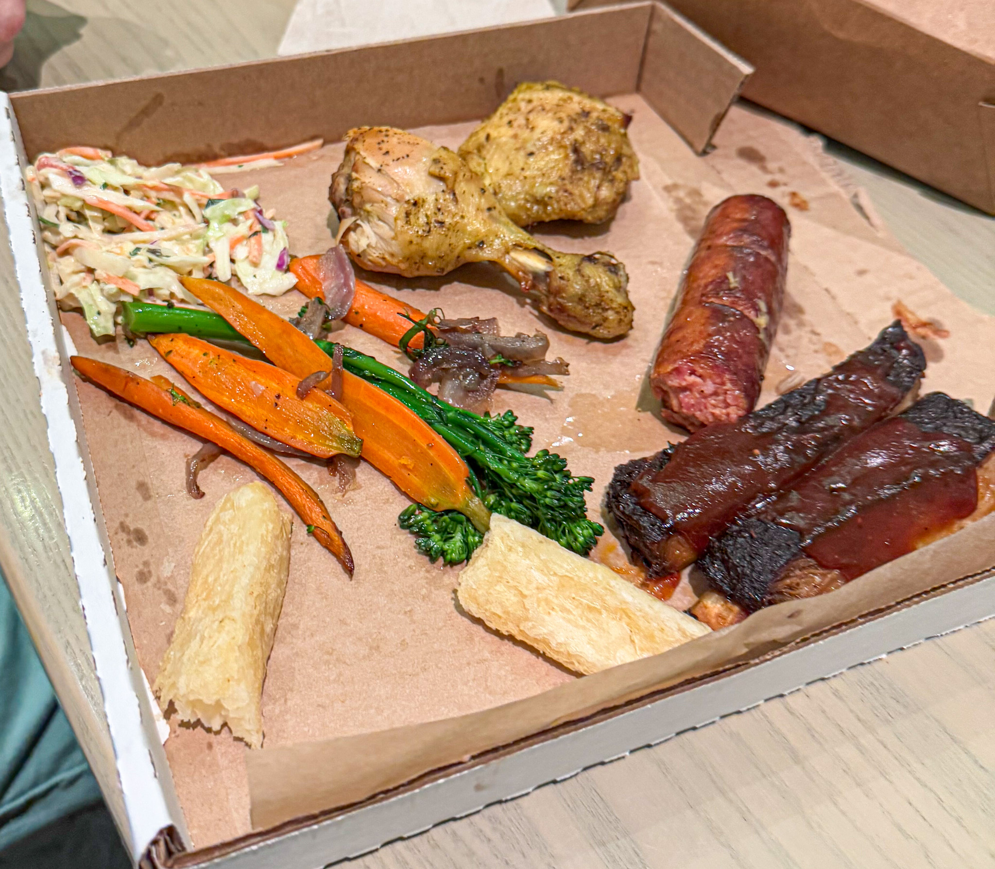A takeout box with coleslaw, roasted vegetables, chicken drumstick, sausage, ribs, and two breadsticks from Centertown Market at Disney.