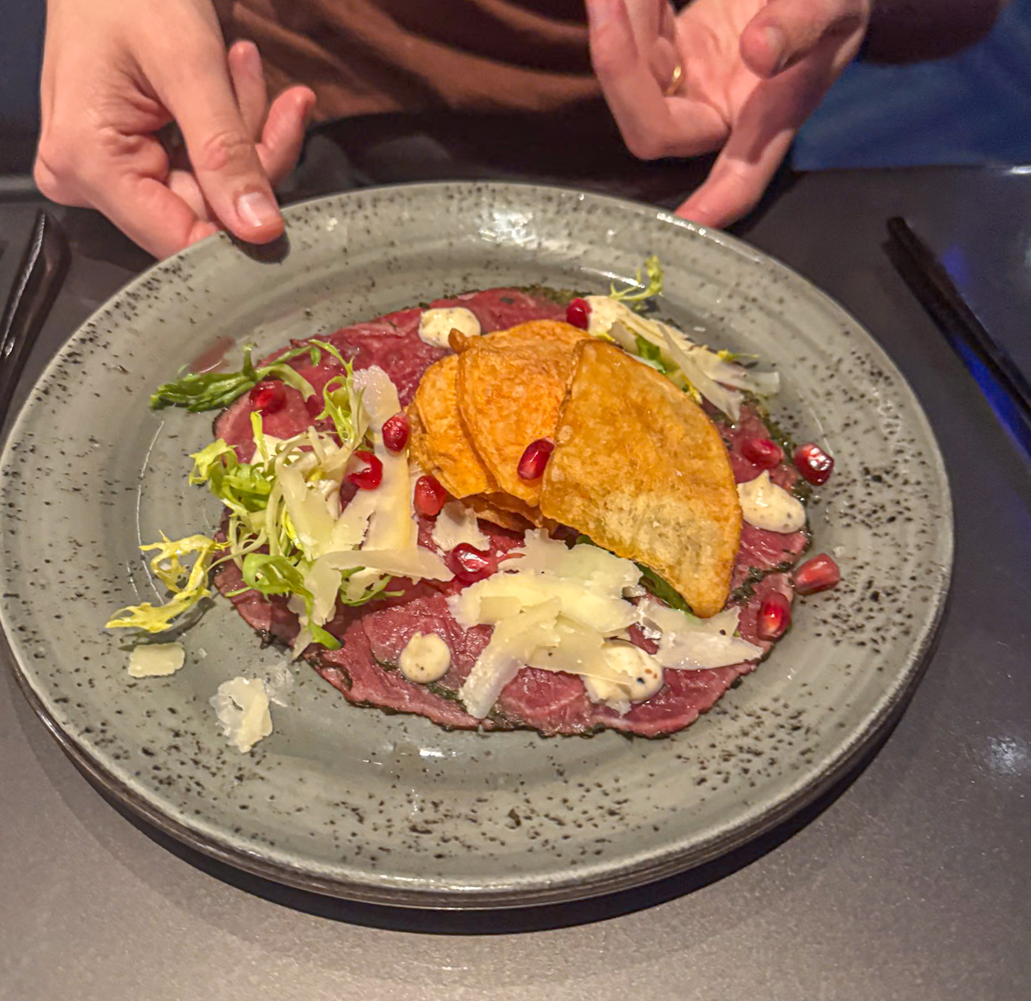 A plate of beef carpaccio topped with cheese, greens, pomegranate seeds, and crispy chips.