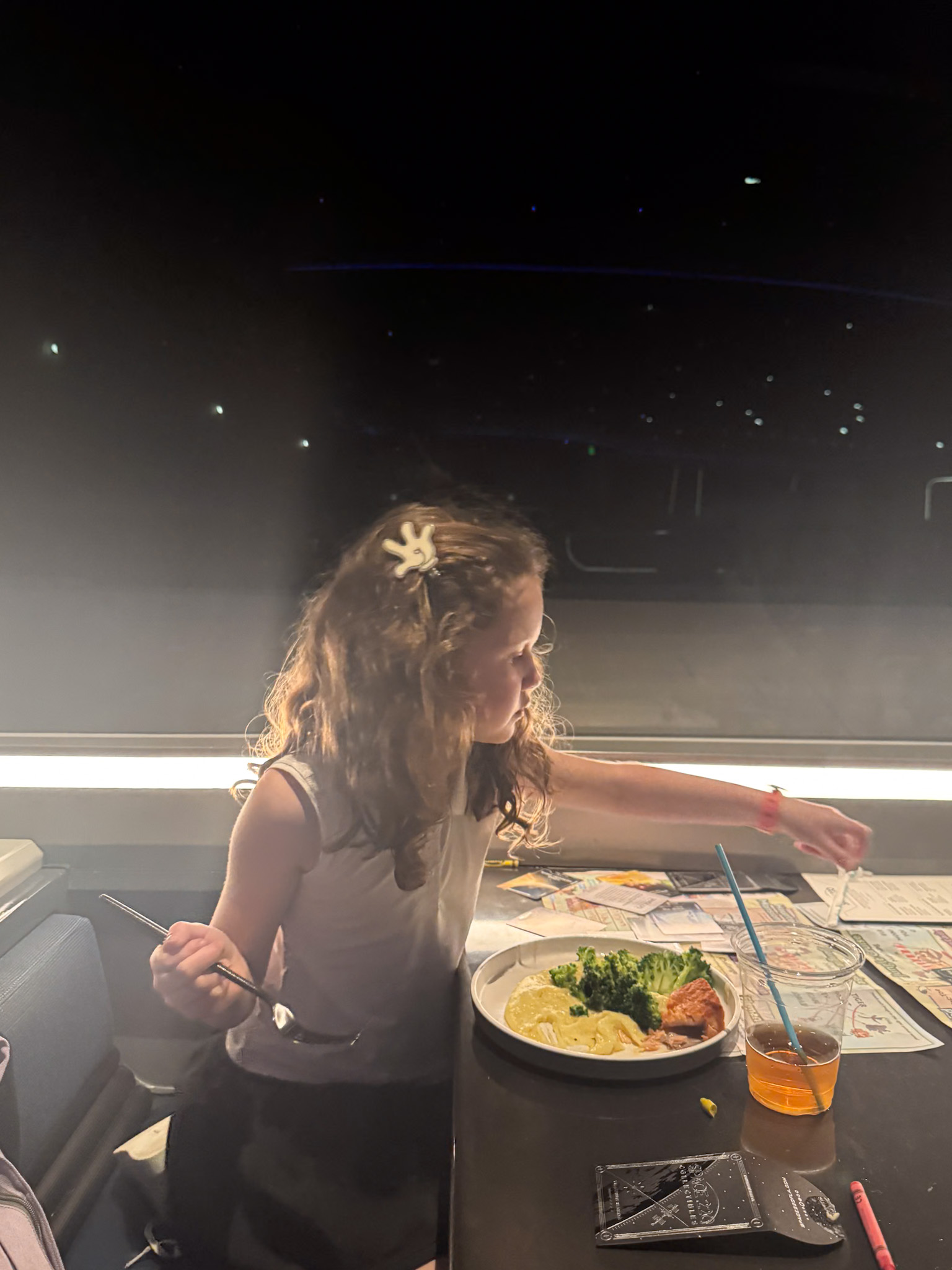 A young girl with curly hair eats dinner at a dimly lit table, reaching for something beside her.