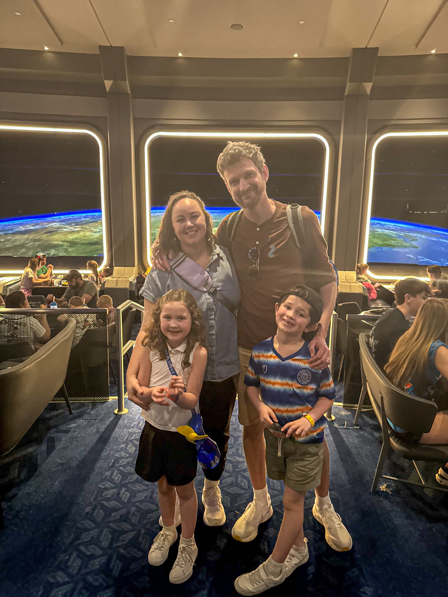 A smiling family of four poses together in front of a large space-themed window display at Disney's Space 220.