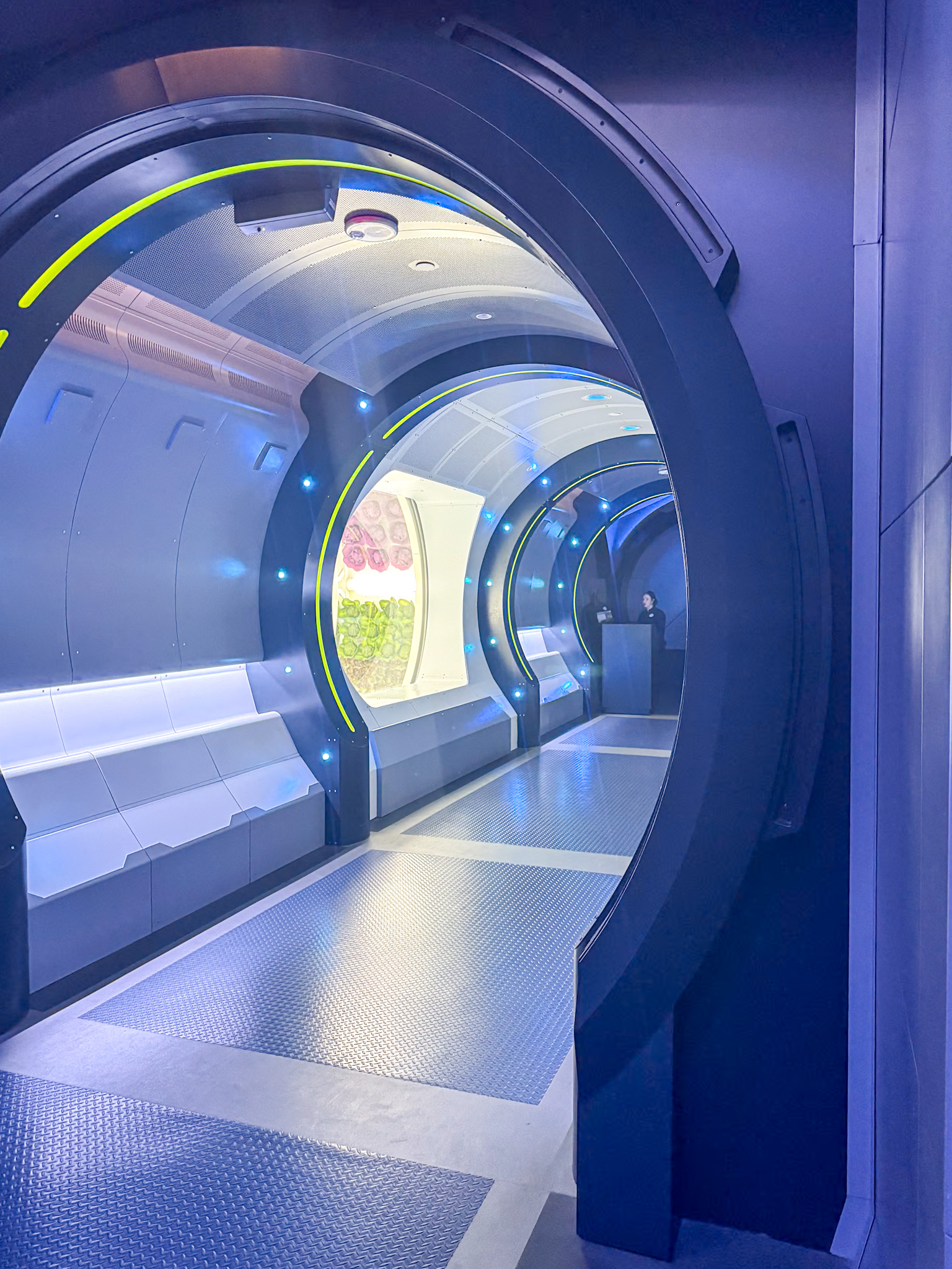 Futuristic hallway with circular doorways, blue lighting, and white benches along the wall at the Space 220 restaurant at Walt Disney World.