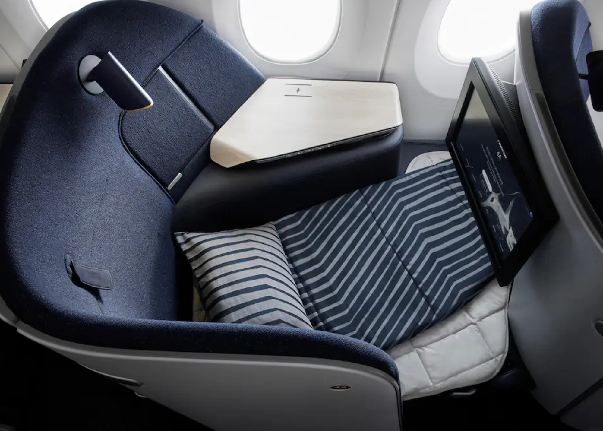 A business class airplane seat reclined flat with a striped blanket and pillow, next to a screen and table.