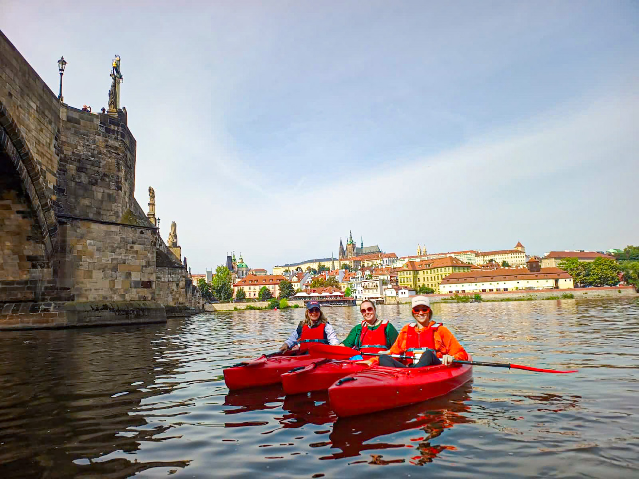 Three people in red kayaks paddle on a river near a historic stone bridge and city buildings in the background.