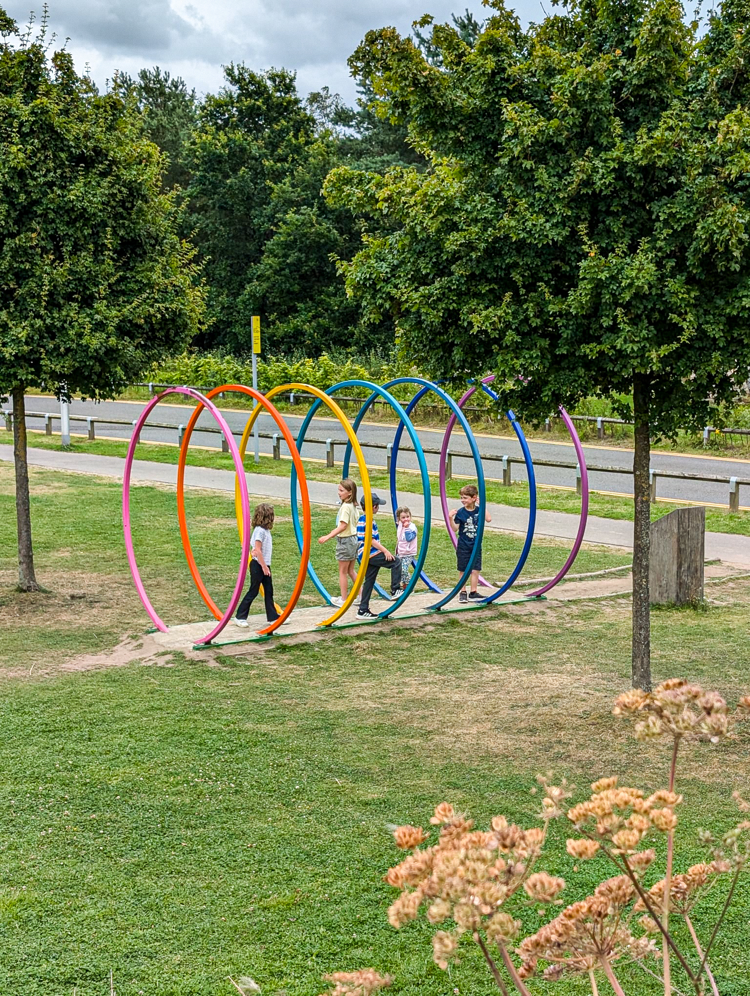 Four children walk through large colorful hoop sculptures in a park with trees and grass.