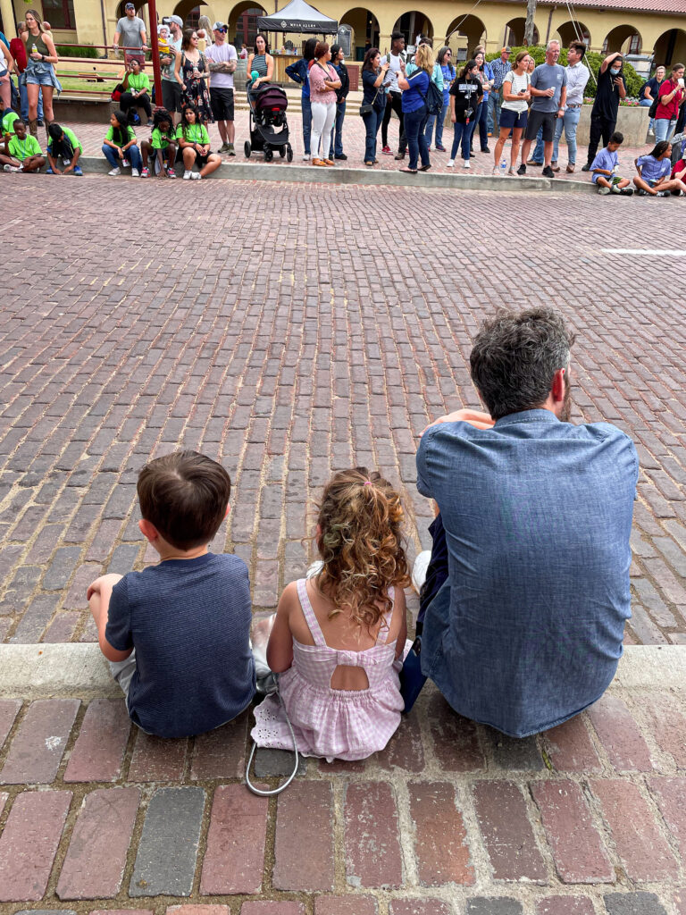 A man and two children sit on a curb, facing a crowd across a cobblestone street.