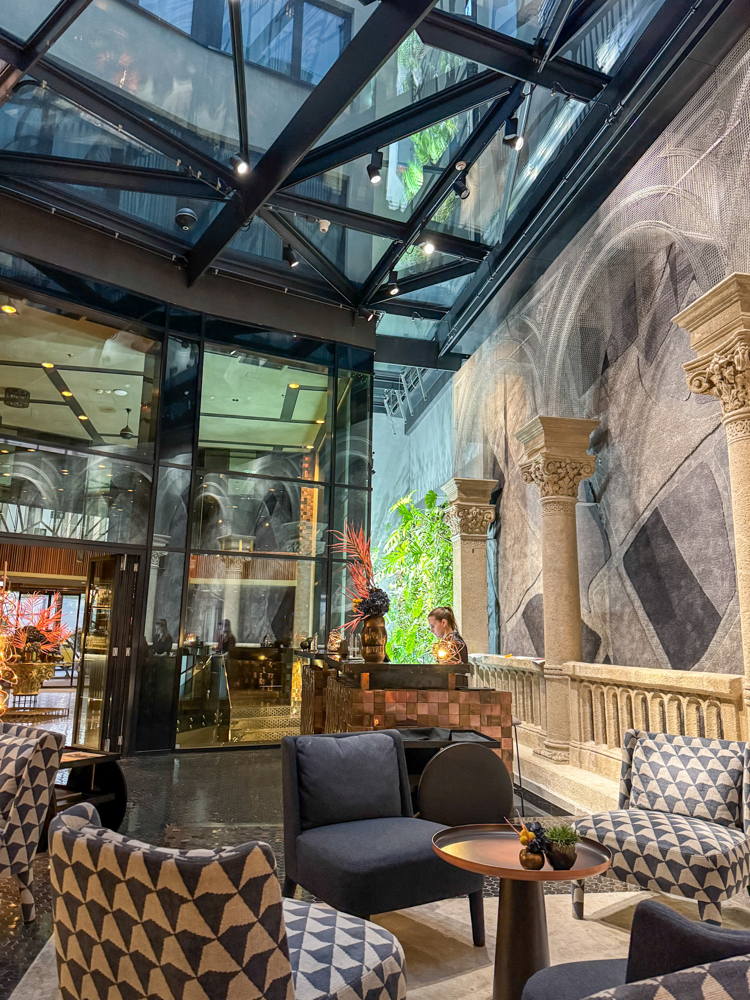 Modern hotel lobby with patterned chairs, stone columns, glass ceiling, and greenery on the wall at the Hotel Clark in Budapest.