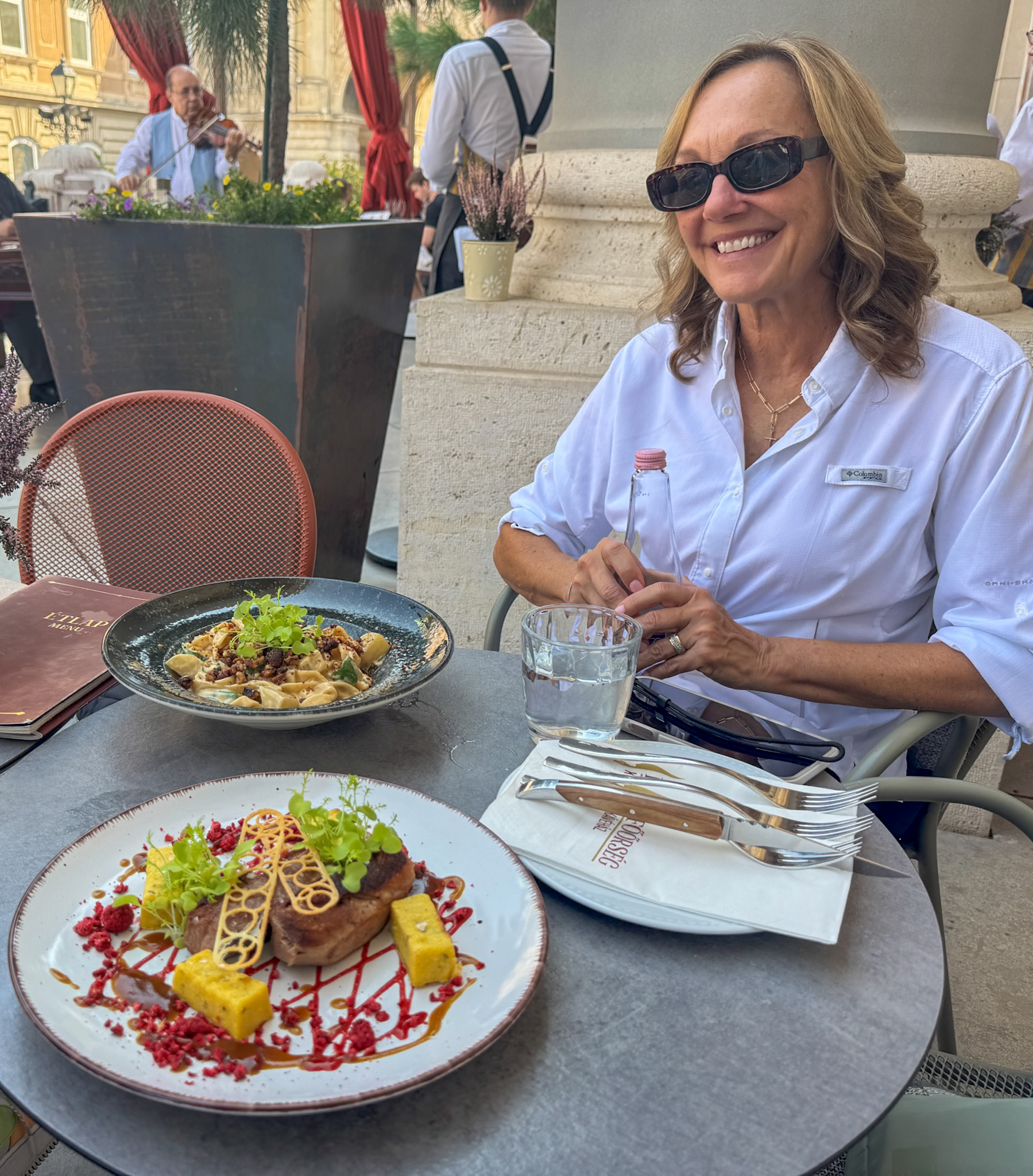 Smiling woman in sunglasses sits at an outdoor café table with gourmet dishes at the Royal Guard Cafe at Buda Castle