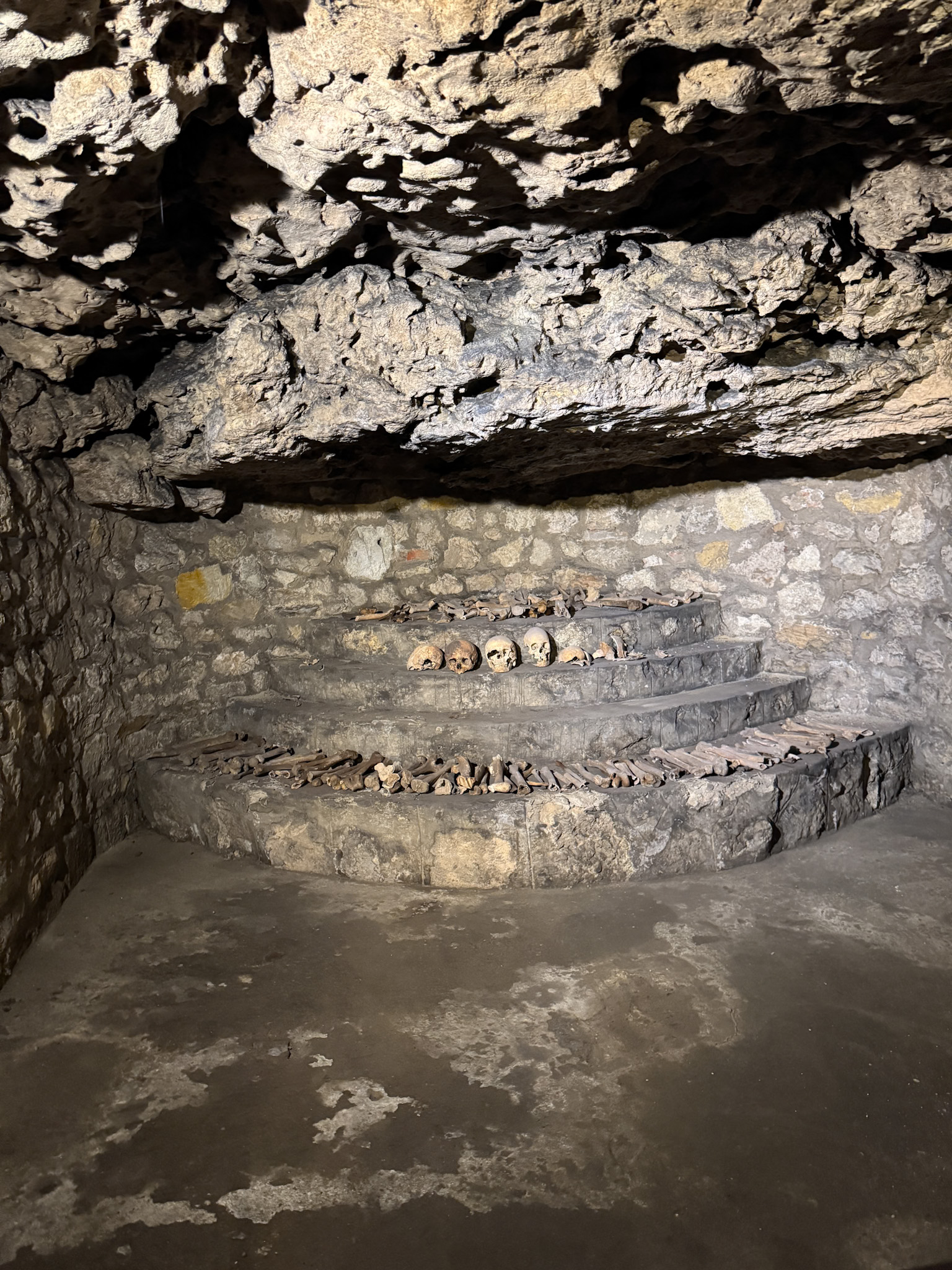 Stone steps in a cave-like chamber, lined with human skulls and bones against a rough stone wall in the Buda Castle Caves.