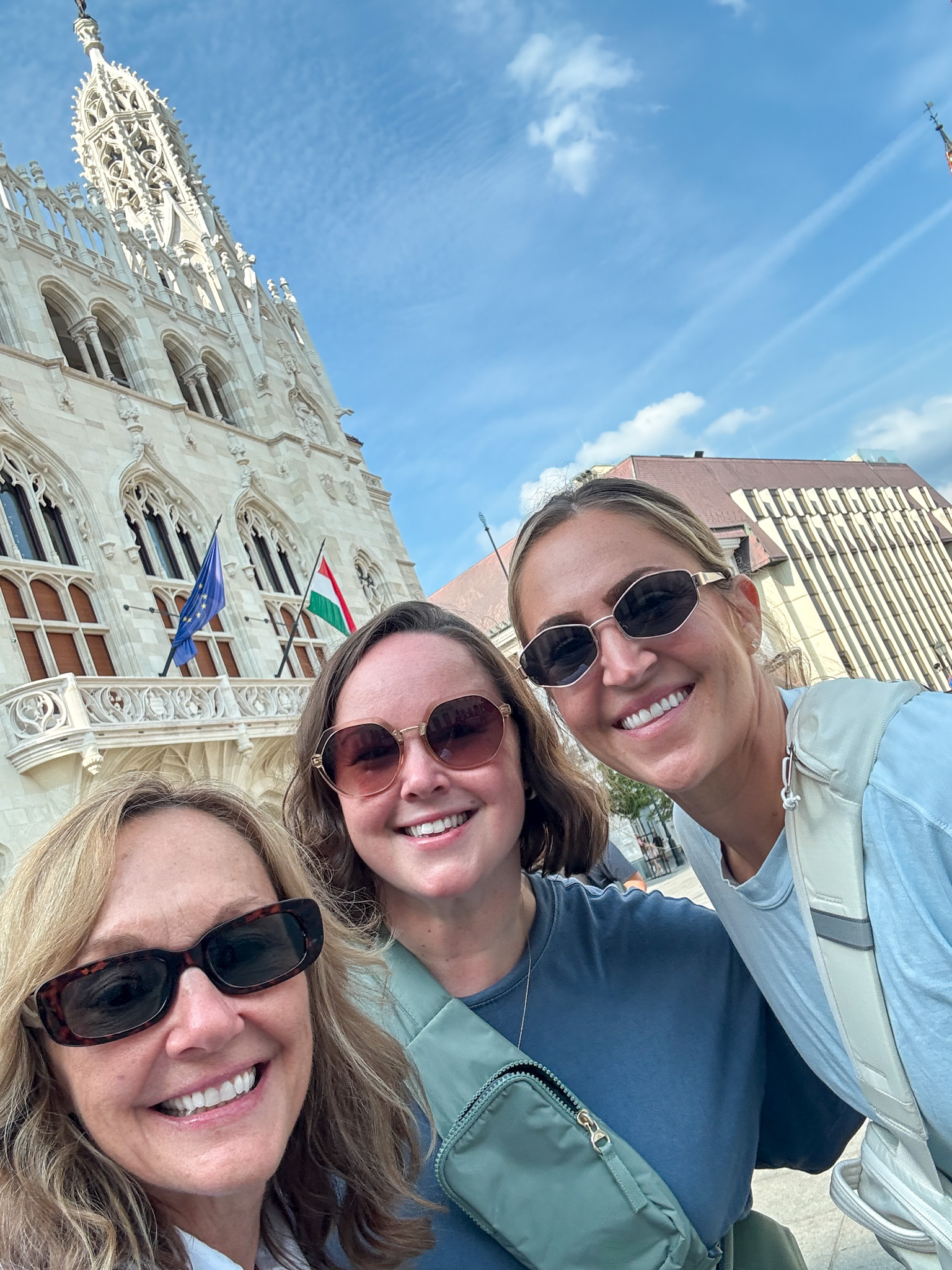Three women in sunglasses smile in front of a historic white building with flags on a sunny day on their 2 day Budapest itinerary.