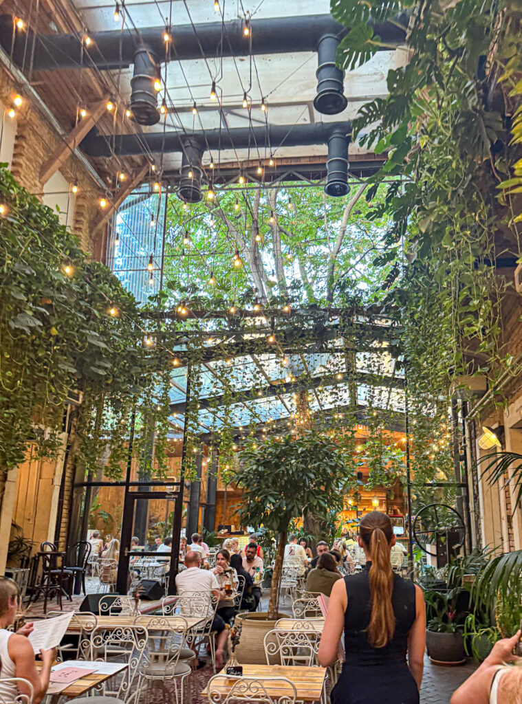 A lush, plant-filled café with string lights, glass ceiling, and people dining at white tables at Mazel Tov.