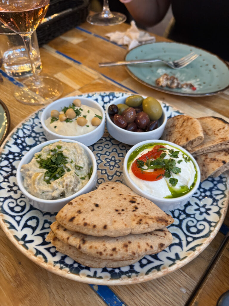 A plate with pita bread, three dips, and olives on a blue patterned dish at a restaurant table at Mazel Tov in Budapest.