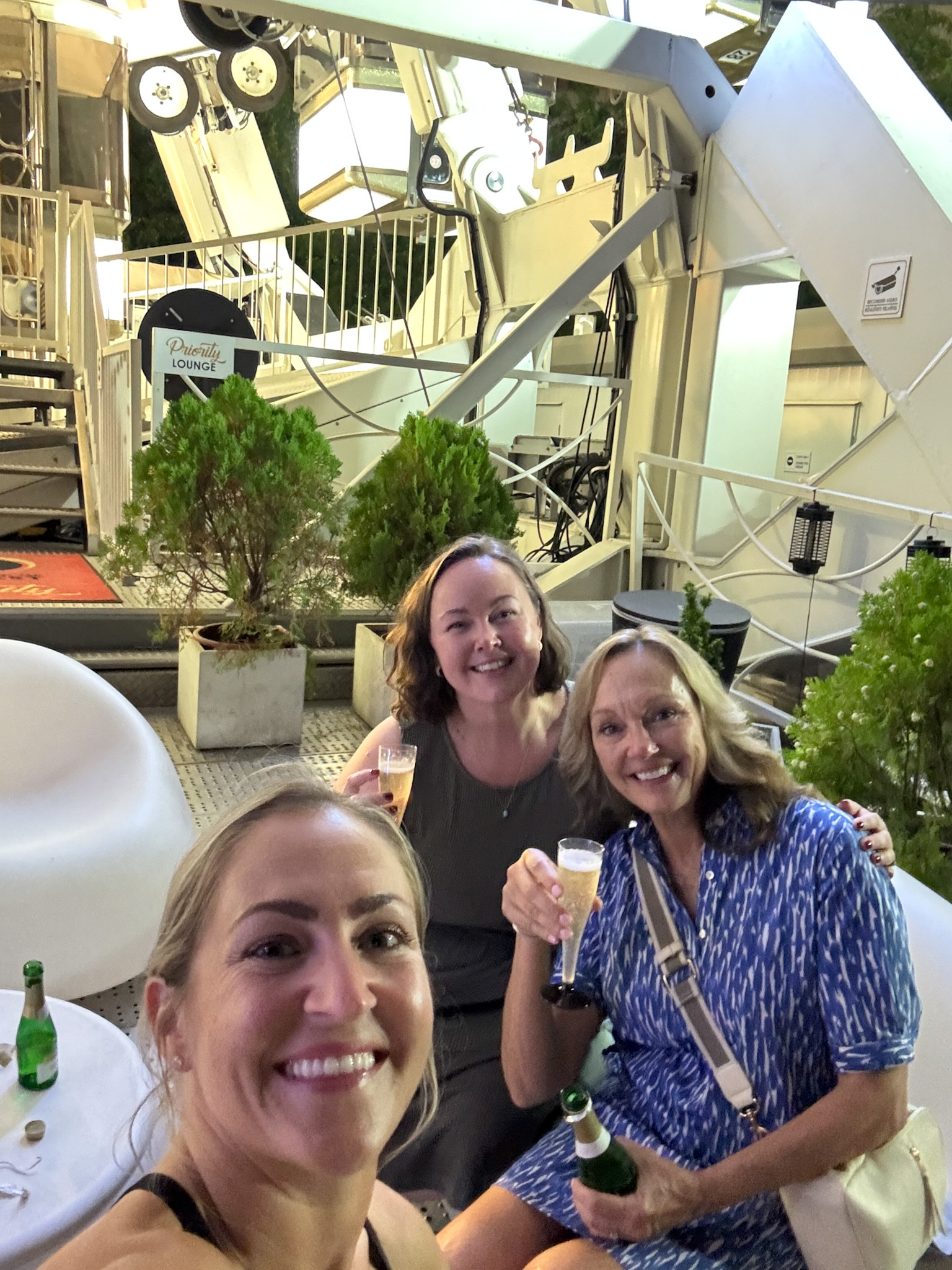 Three women smiling and holding drinks outside the Ferris Wheel of Budapest.