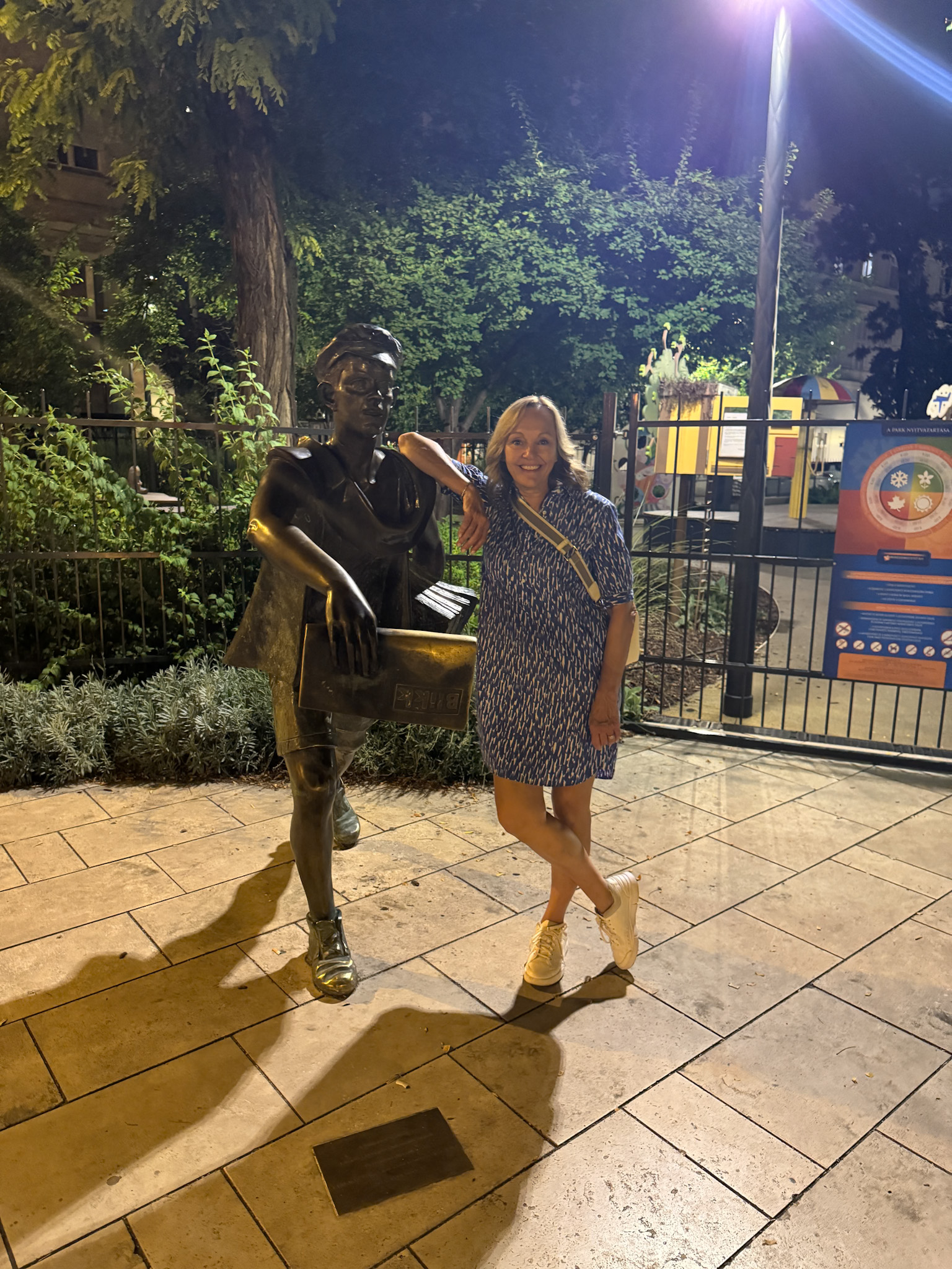 A smiling woman poses at night, leaning on a bronze statue of a person holding a book in a park in Budapest.